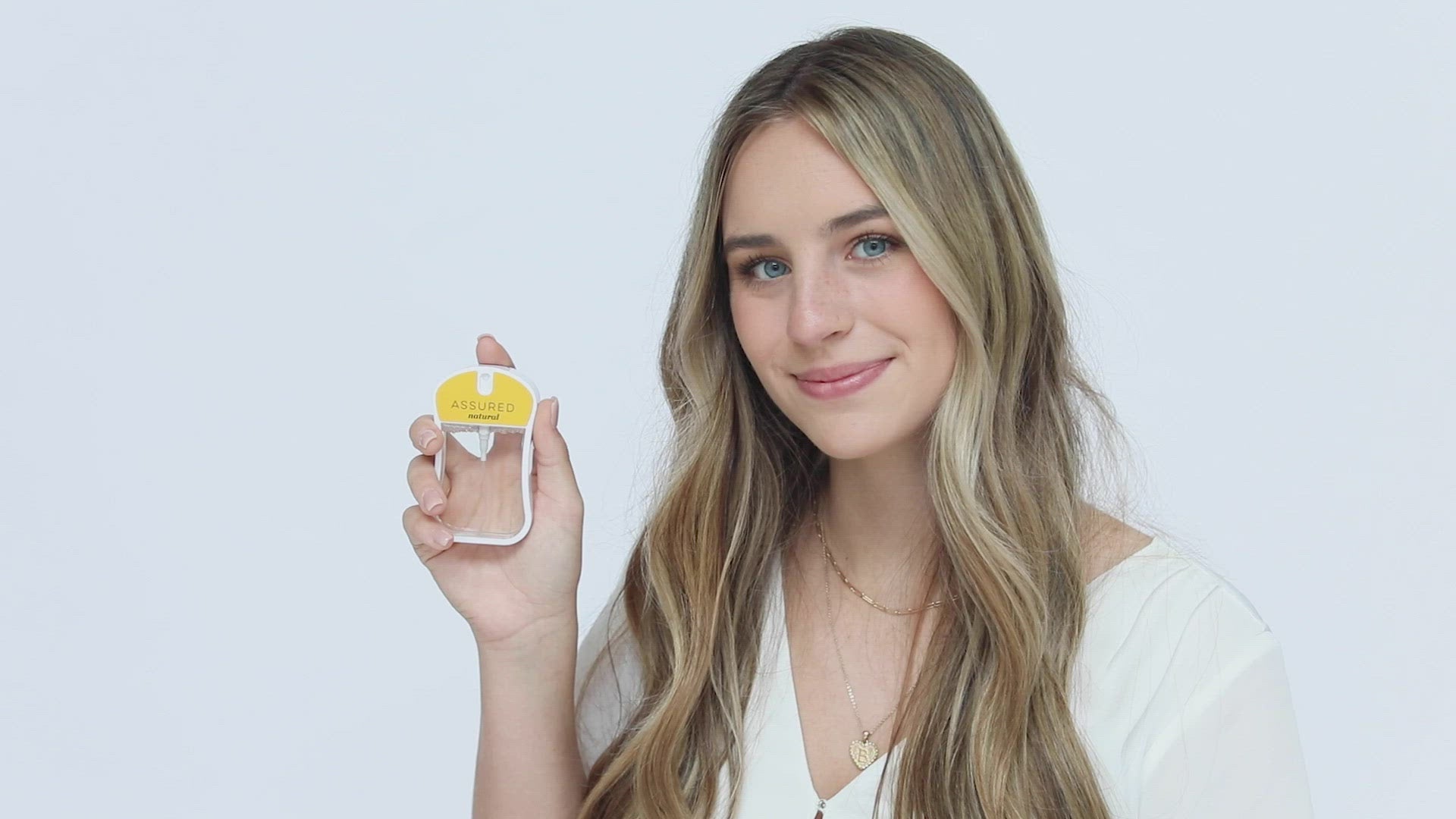 Assured (Natura) Moisturizing Body Mist in mustard color box and rounded rectangle spray bottle with mustard color label and clear liquid. Spray bottle has 1.1 fl oz or 35 mL. Spray bottle and box are pictured with model holding spray bottle in her hand and spraying it then smelling it and smiling.