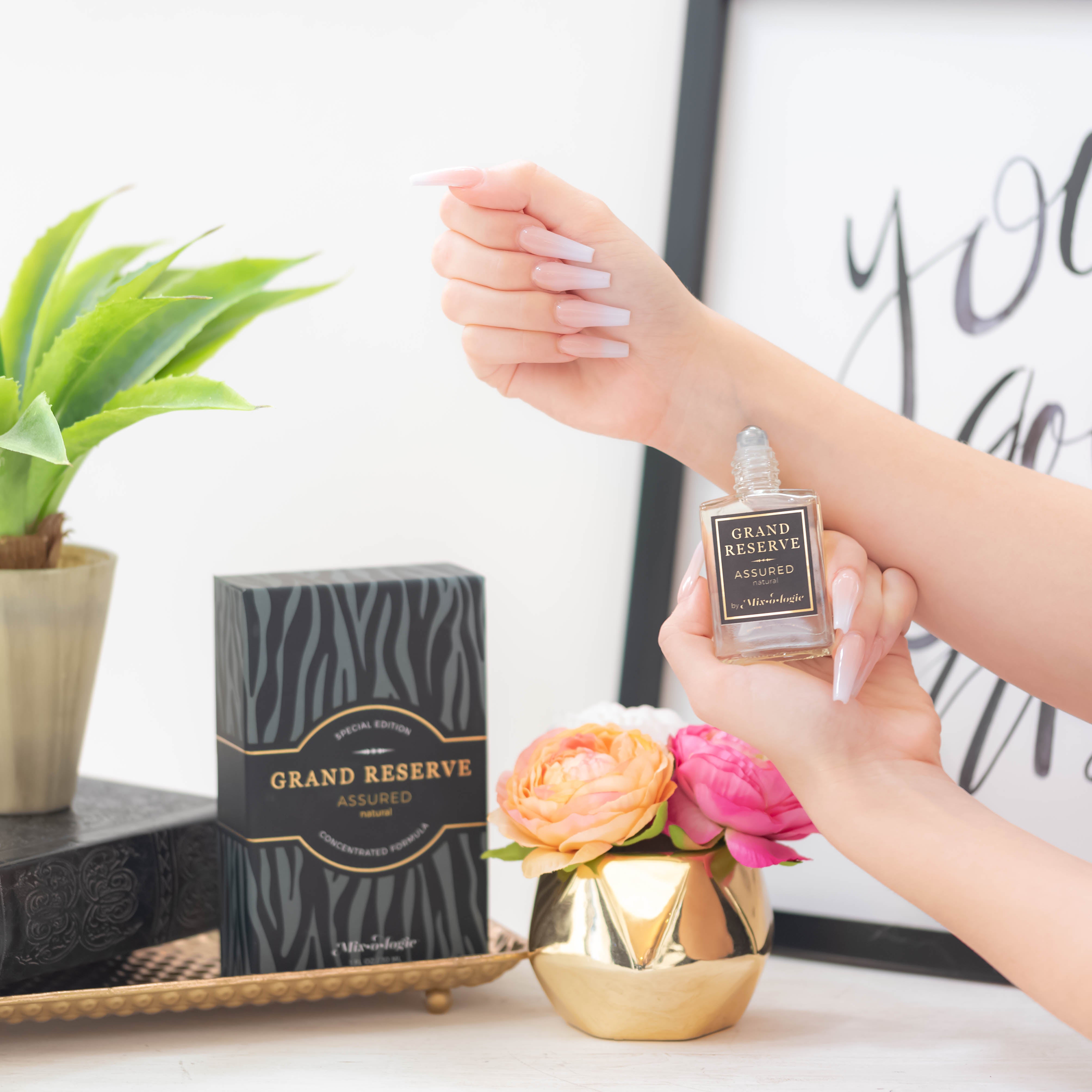 Assured (Natural) Special Edition Grand Reserve concentrated formula in clear glass rectangle perfume bottle that has 1 fl oz or 30 mL of light-yellow liquid with black cap, label, and spray nozzle. Black zebra pattern rectangle box with mustard letting. Grand Reserve and box pictured being applied to wrist with plant, gold vase with flowers. 