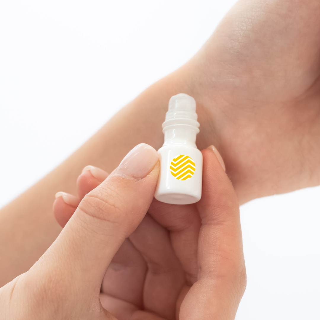 Assured (Natural) mini white cylinder rollerball with mustard chevron pattern has .035 fl oz or 1 mL.  Roll-on perfume oil. Mini rollerball being applied to wrist by model. 