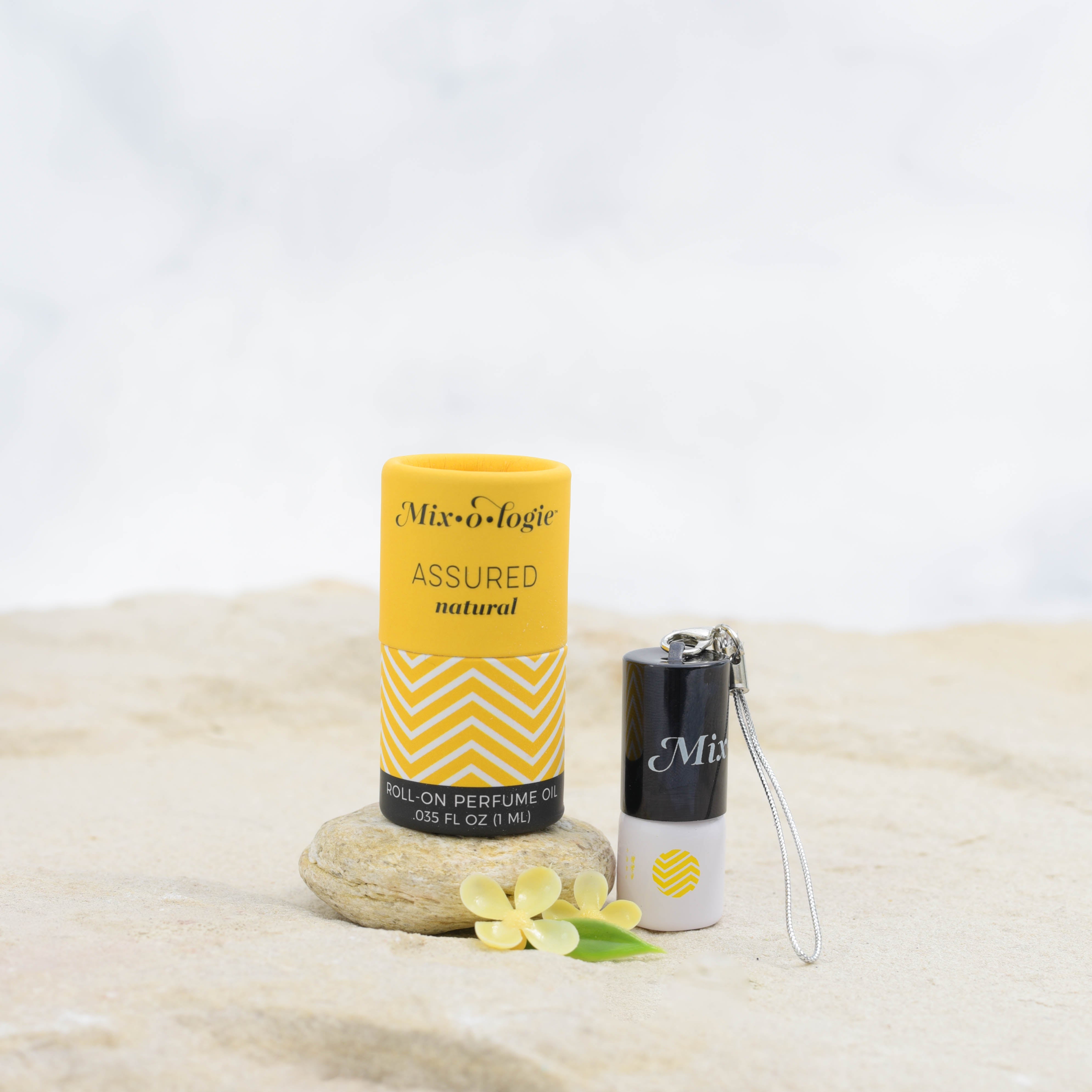 Assured (Natural) mini white cylinder rollerballs with black top and keychain attachment with mustard chevron pattern has .035 fl oz or 1 mL. Mustard cylinder packing tube with mustard chevron pattern. Roll-on perfume oil. Mini rollerball and cylinder tube are in sand on a rock with yellow flowers.