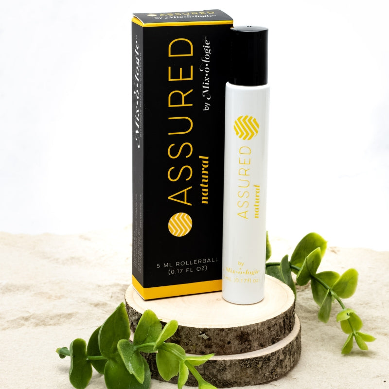 Assured (Natural) white cylinder rollerball with mustard color lettering with black box and mustard color lettering. Rollerball has 0.17 fl oz or 5 mL. Rollerball and rollerball box pictured on wood in the sand with greenery.  