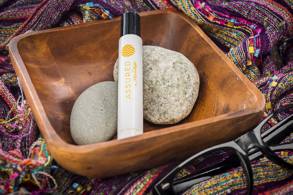 Assured (Natural) white cylinder rollerball with mustard color lettering. Rollerball has 0.17 fl oz or 5 mL. Rollerball pictured in wood container with rocks on a colorful blanket. 