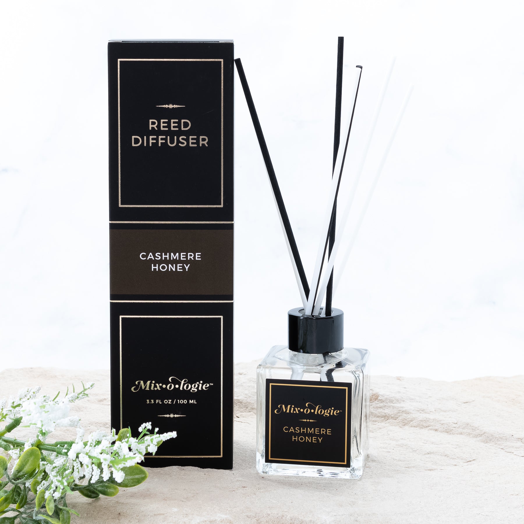 Cashmere Honey Reed Diffuser is a clear square glass container with black label that says Mixologie – Cashmere Honey. Has a black top and 4 white & 4 black reed sticks coming out of top, is 3.3 fl oz or 100 mL of clear scented liquid. Black rectangle package box with brown label. Box and diffuser are pictured with sand and greenery with white flowers.