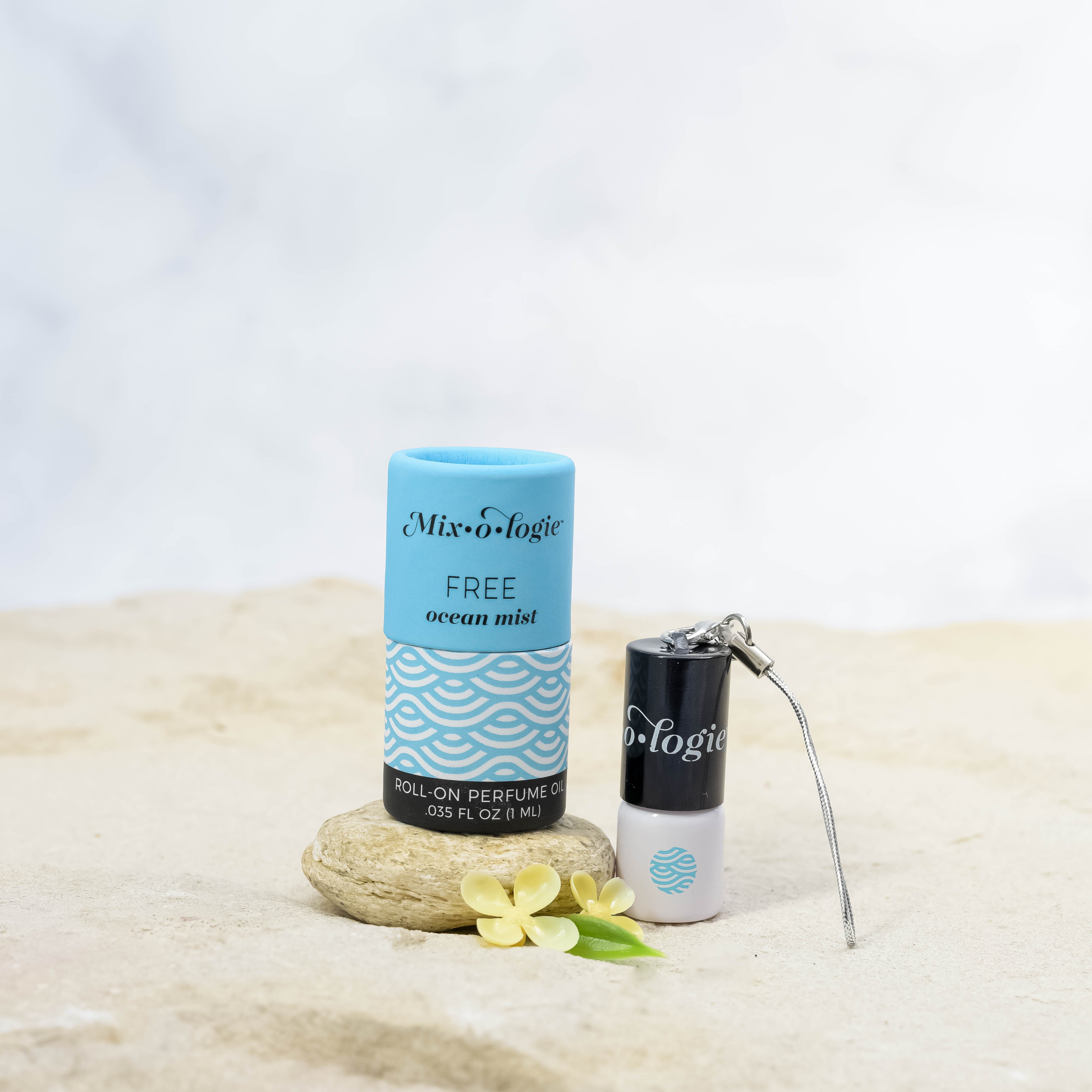 Free (Ocean Mist) mini white cylinder rollerballs with black top and keychain attachment with pale blue wave-like pattern and has .035 fl oz or 1 mL. Pale blue cylinder packing tube with pale blue wave-like pattern. Roll-on perfume oil. Mini rollerball and cylinder tube are in sand on a rock with yellow flowers.