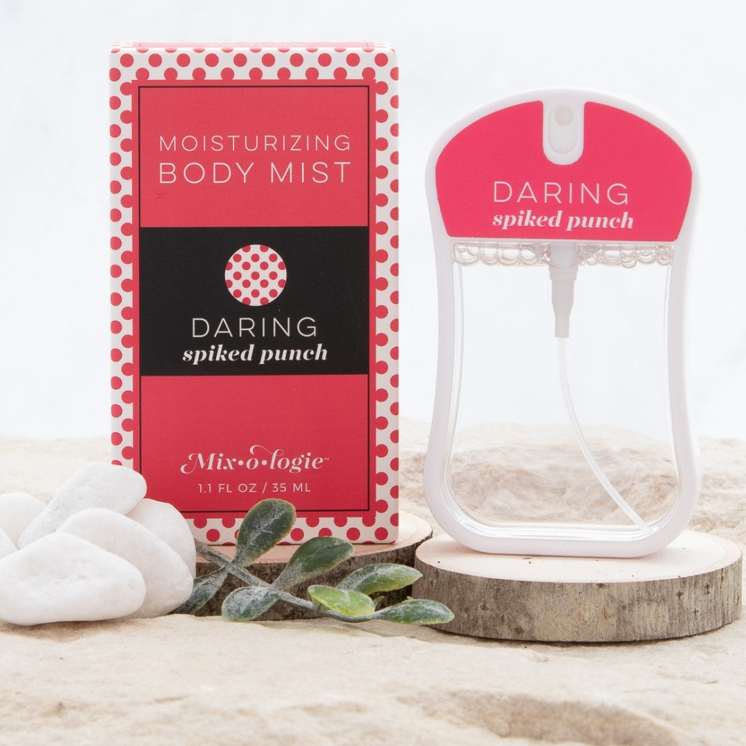 Daring (Spiked Punch) Moisturizing Body Mist in bright pink color box and rounded rectangle spray bottle with bright pink color label and clear liquid. Spray bottle has 1.1 fl oz or 35 ML. Spray bottle and box are pictured in the sand on wood with pebbles and greenery.