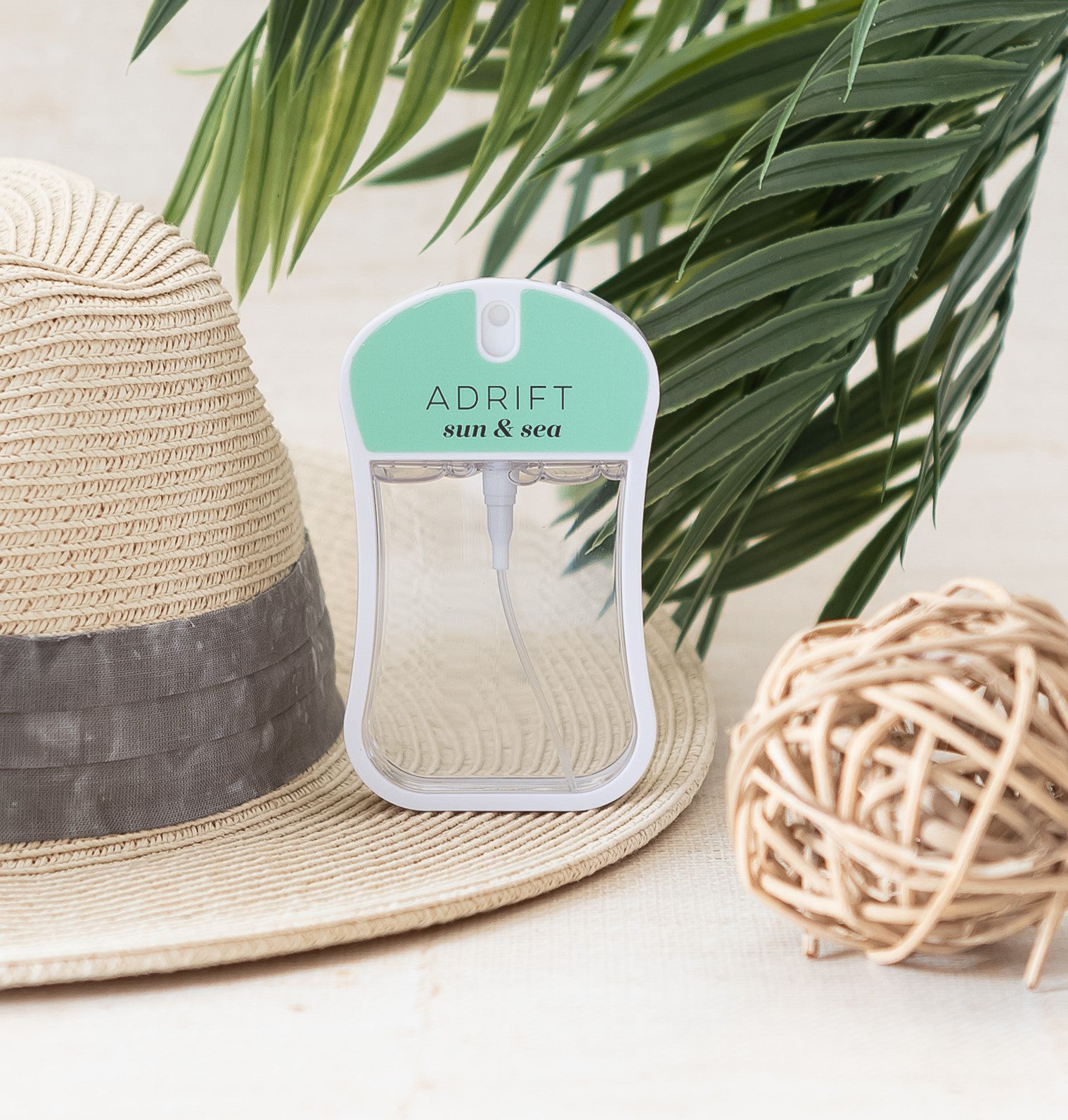 Adrift (Sun & Sea) moisturizing body mist spray bottle with light green color and clear liquid in a rounded rectangle shape. Spray bottle has 1.1 fl ox or 35 mL. spray bottle is on straw hat with palm tree in background.