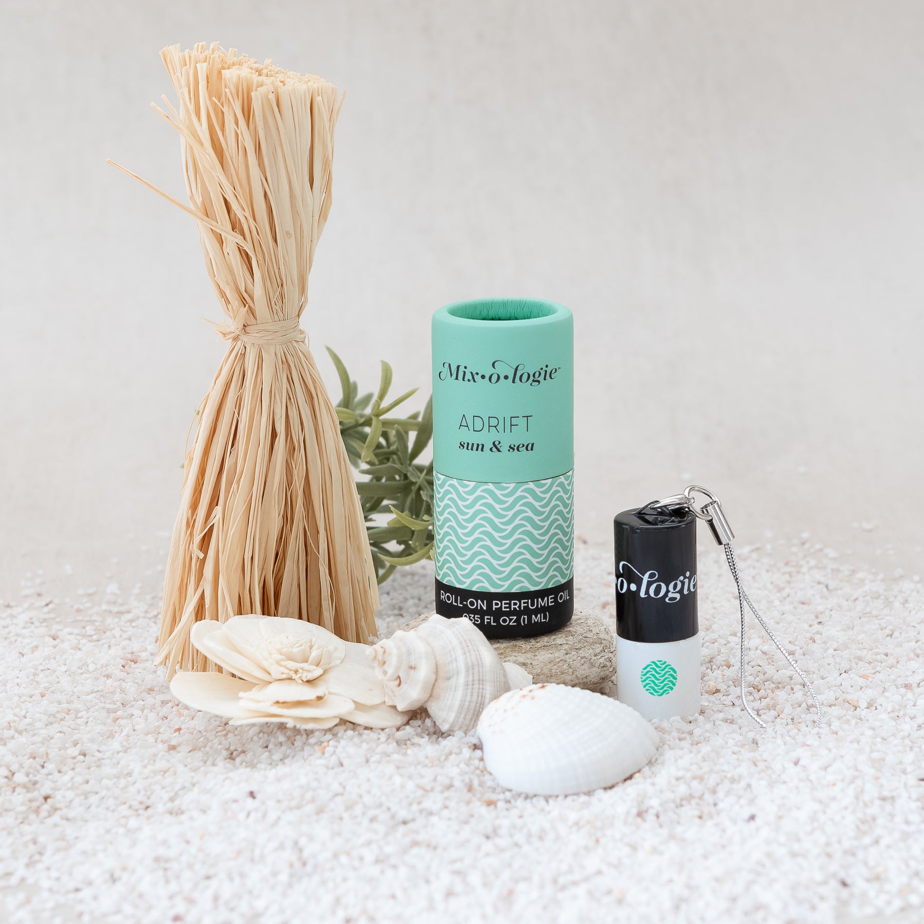 Adrift (Sun & Sea) mini white cylinder rollerballs with black top and keychain attachment with light green Adrift pattern has .035 fl oz or 1 mL. Light green cylinder packing tube. Mini rollerball and cylinder tube are in sand on rock with sea shells, straw, and greenery.