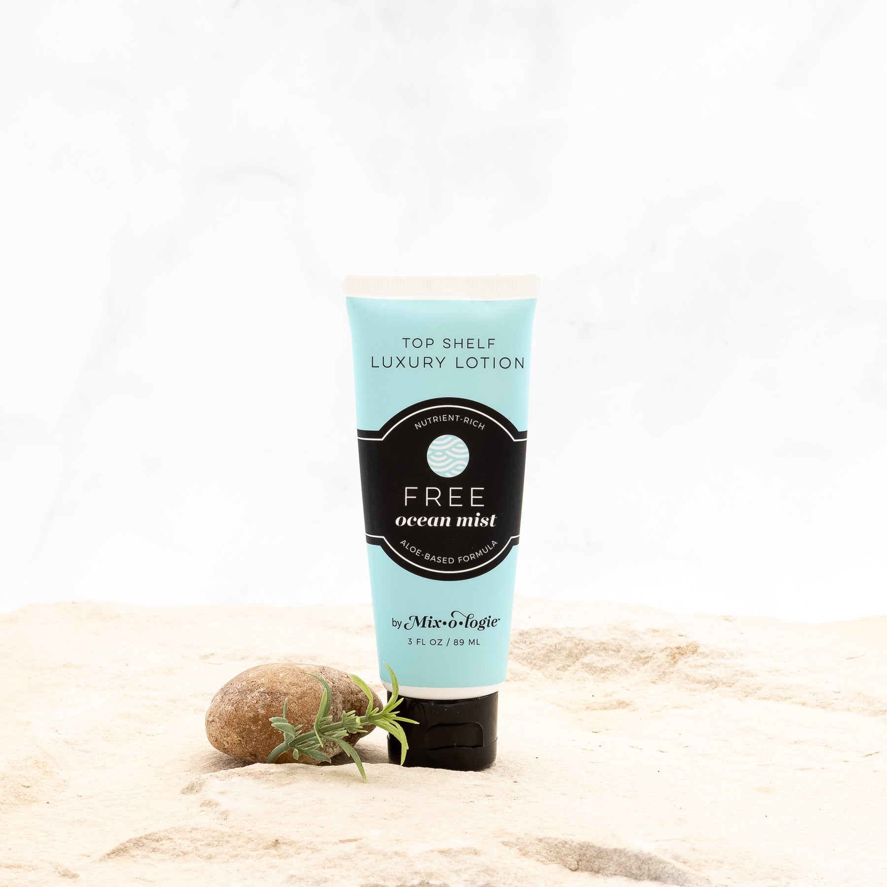 Free (Ocean Mist) Top Shelf Lotion in pale blue tube with black lid and label. Nutrient rich, aloe-based formula, tube has 5 fl oz or 89 mL. Pictured with white background, sand, rocks, and greenery. 
