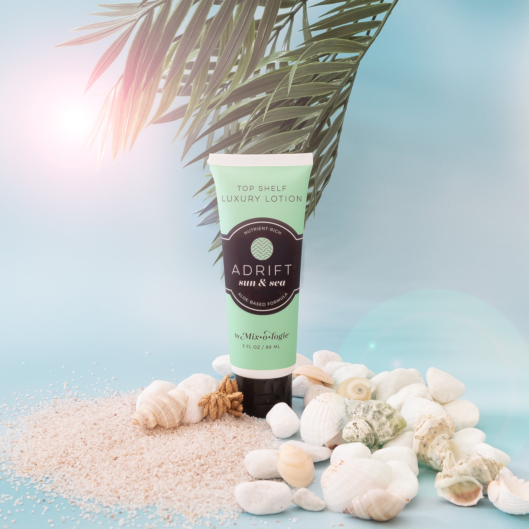 Adrift (Sun & Sea) Top shelf lotion in light green tube with black lid and label. Nutrient rich, aloe based formula, tube has 5 fl oz or 89 mL. Pictured with blue background, sand, seashells and green palm tree.