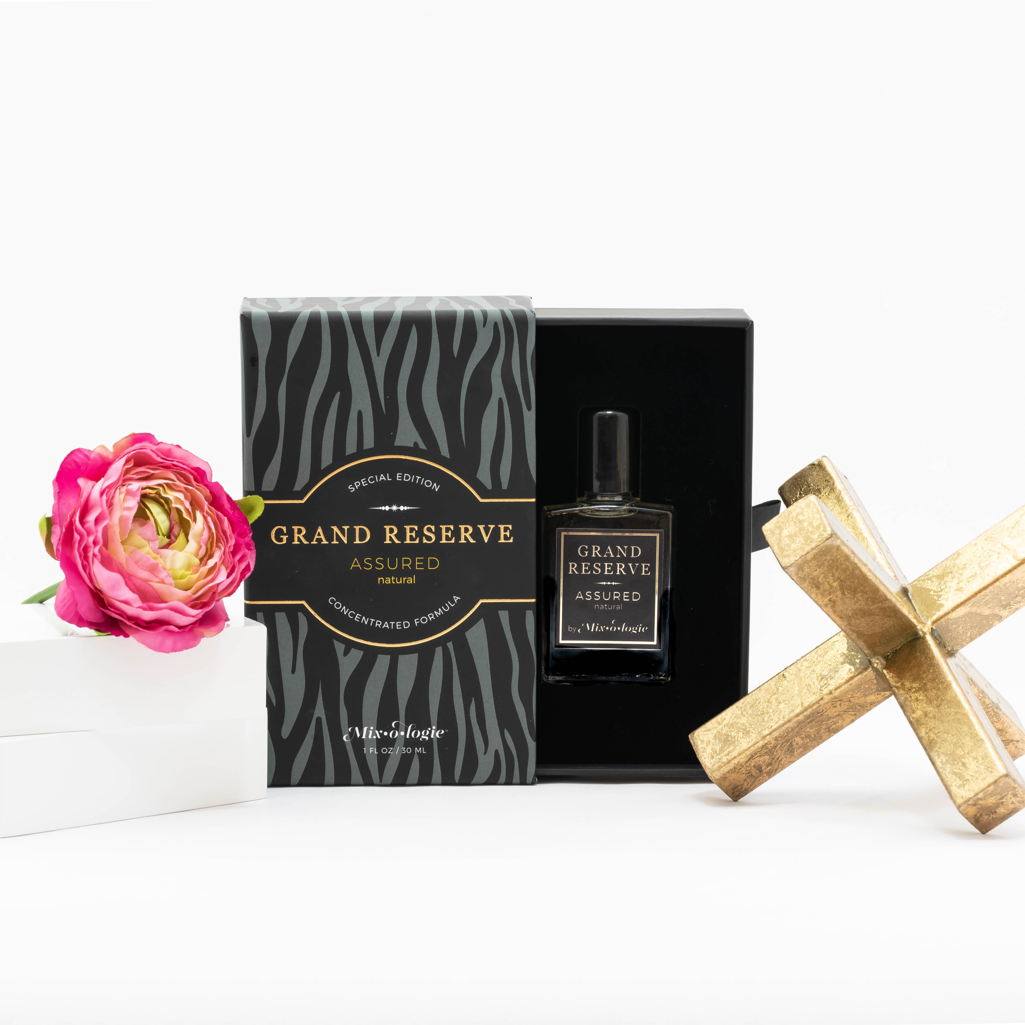 Assured (Natural) Special Edition Grand Reserve concentrated formula in clear glass rectangle perfume bottle that has 1 fl oz or 30 mL of light-yellow liquid with black cap, label, and spray nozzle. Black zebra pattern rectangle box with mustard letting. Grand Reserve and box pictured with white background pink flower and gold table decor