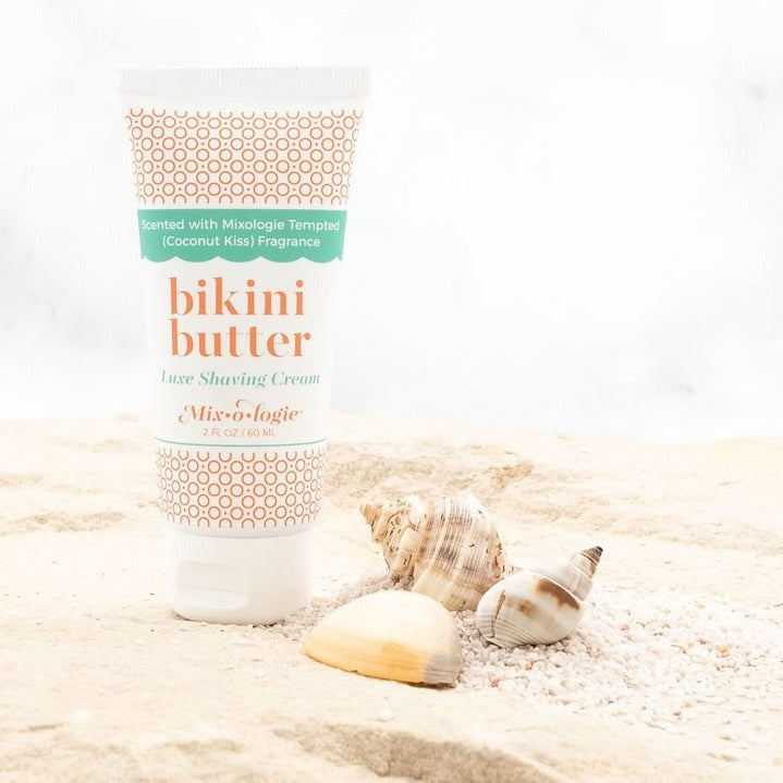 Luxe Shaving Cream Bikini Cutter scented with Mixologie Tempted (Coconut Kiss) Fragrance in a 2 fl oz or 60 mL white tube with orange circle pattern and white cap. Bikini Butter pictured in sand with sea shells. 