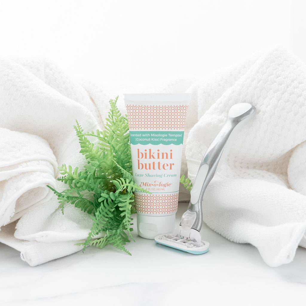 Luxe Shaving Cream Bikini Cutter scented with Mixologie Tempted (Coconut Kiss) Fragrance in a 2 fl oz or 60 mL white tube with orange circle pattern and white cap. Bikini Butter pictured on a white towel with razor and greenery.