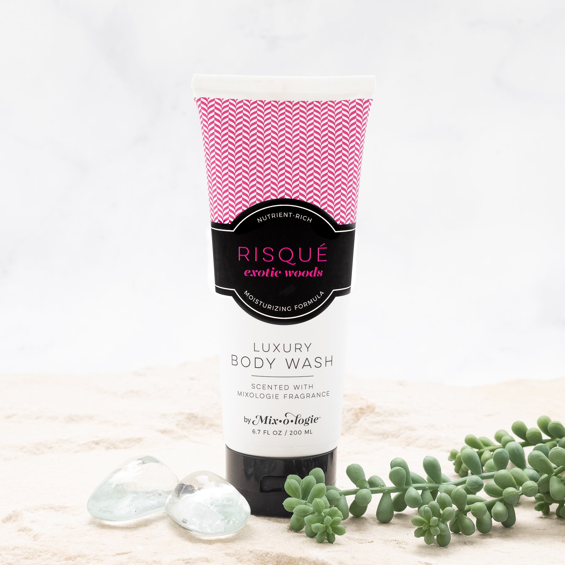 Luxury Body Wash in Risqué (Exotic Woods) in dark pink pattern package and black label in sand with rocks and greenery.