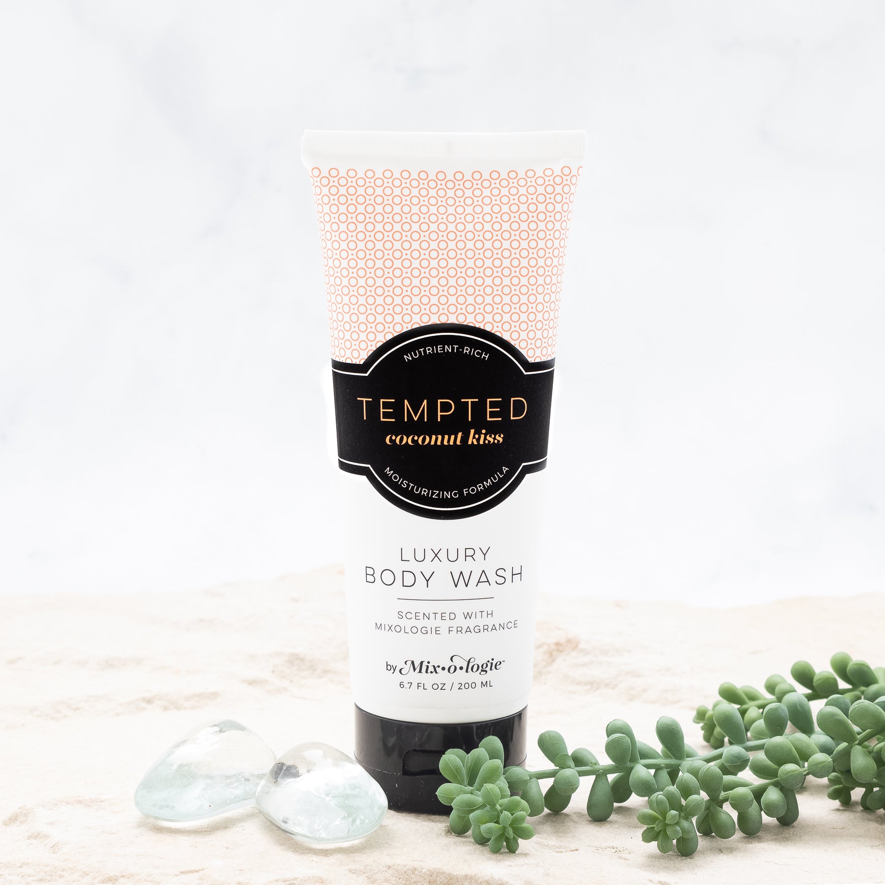 Luxury Body Wash in Mixologie’s Tempted (Coconut Kiss) in peach color sample package with black label. Contains 6.7 fl oz or 200 mL pictured in sand with rocks and greenery.