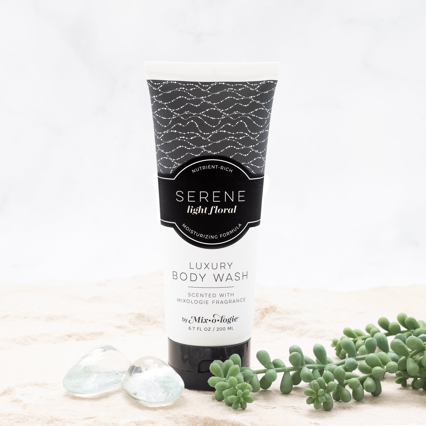 Luxury Body Wash in Serene (Light Floral) in white pattern package and black label in the sand with rocks and greenery .Contains 6.7 fl oz or 200 mL. 