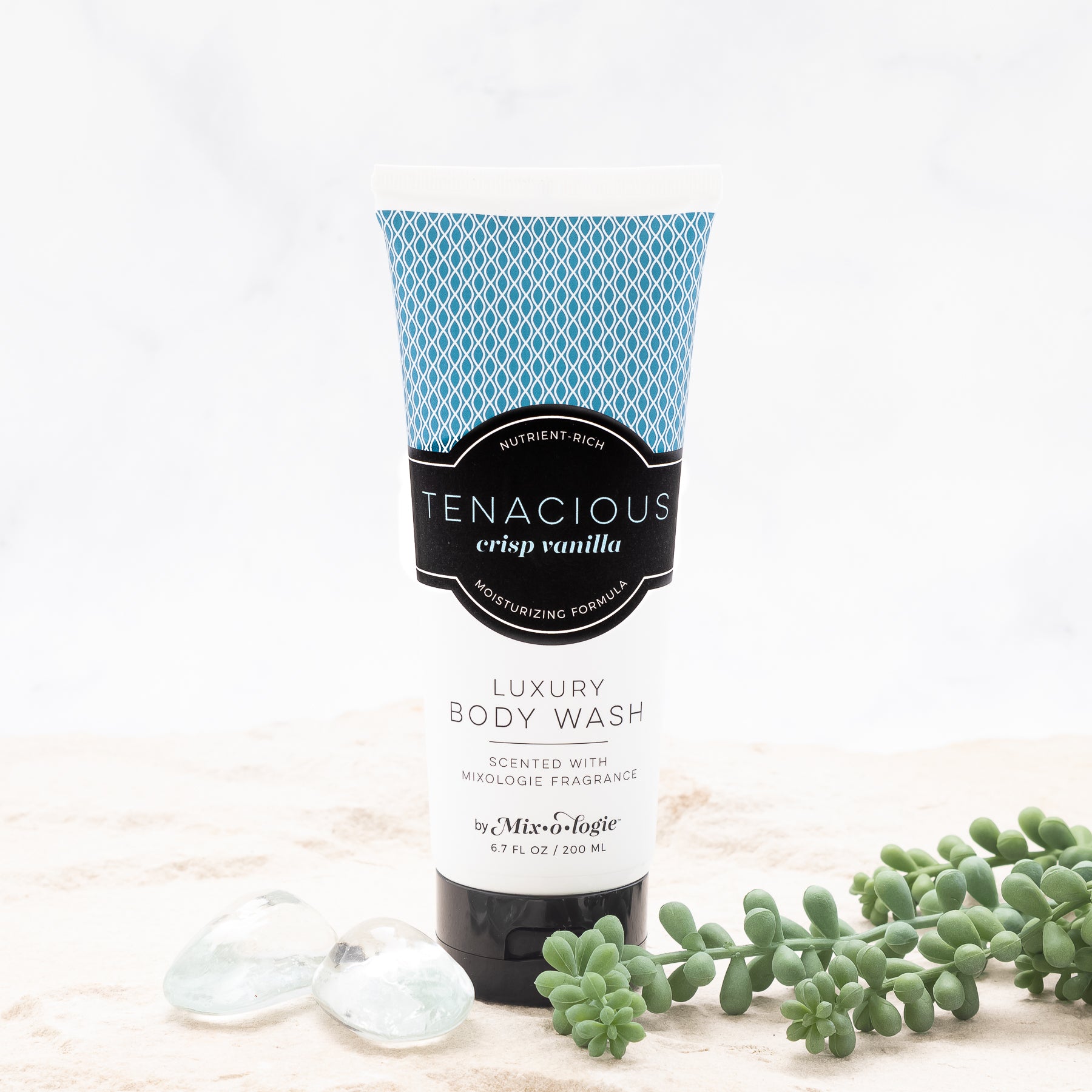 Luxury Body Wash in Mixologie’s Tenacious (Crisp Vanilla) in blue color sample package with black label. Contains 6.7 fl oz or 200 mL pictured in sand with rocks and greenery. 