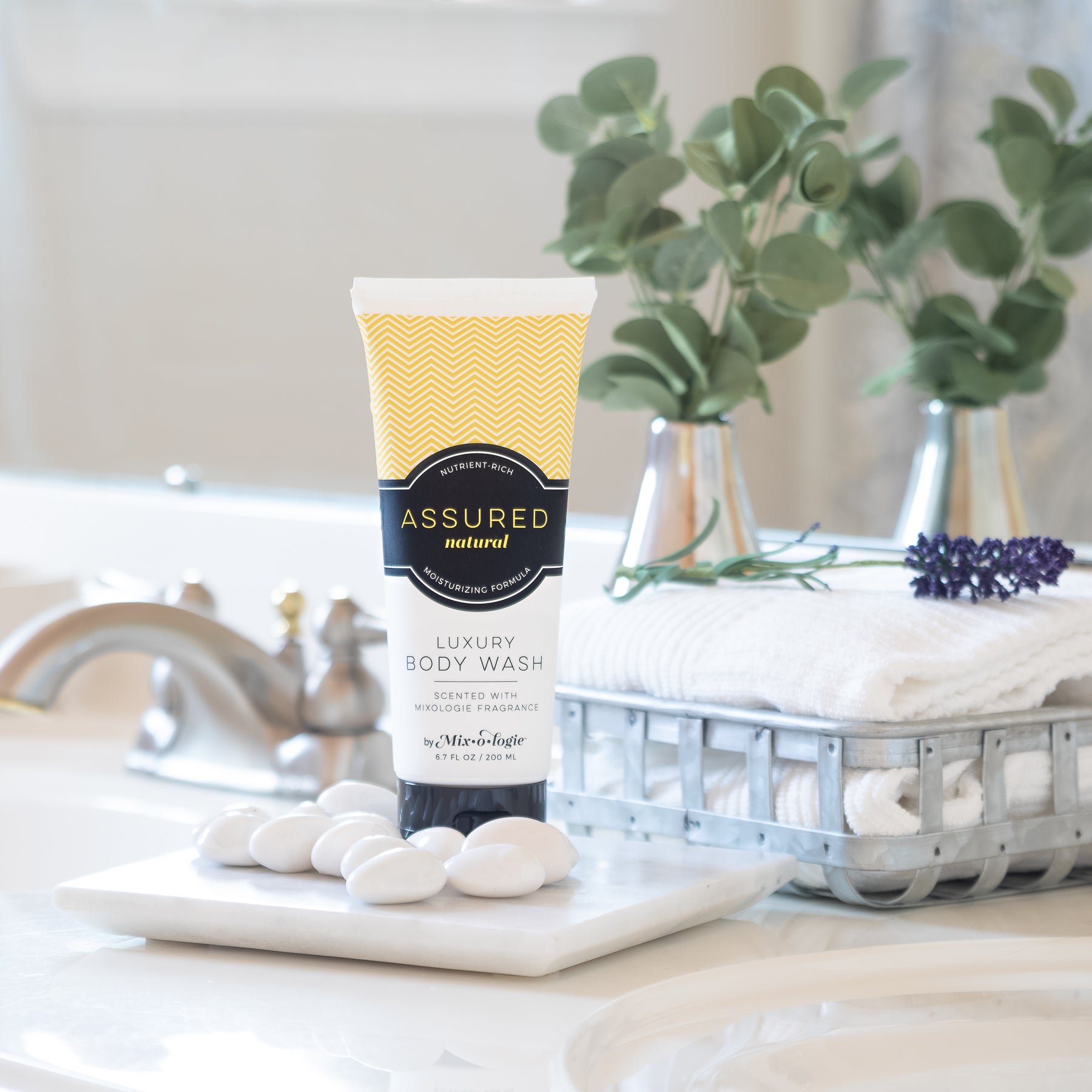 Luxury Body Wash in Mixologie’s Assured (Natural) in yellow color sample package with black label. Contains 6.7 fl oz or 200 mL pictured on a bathroom counter with towels, greenery, and white pebbles.