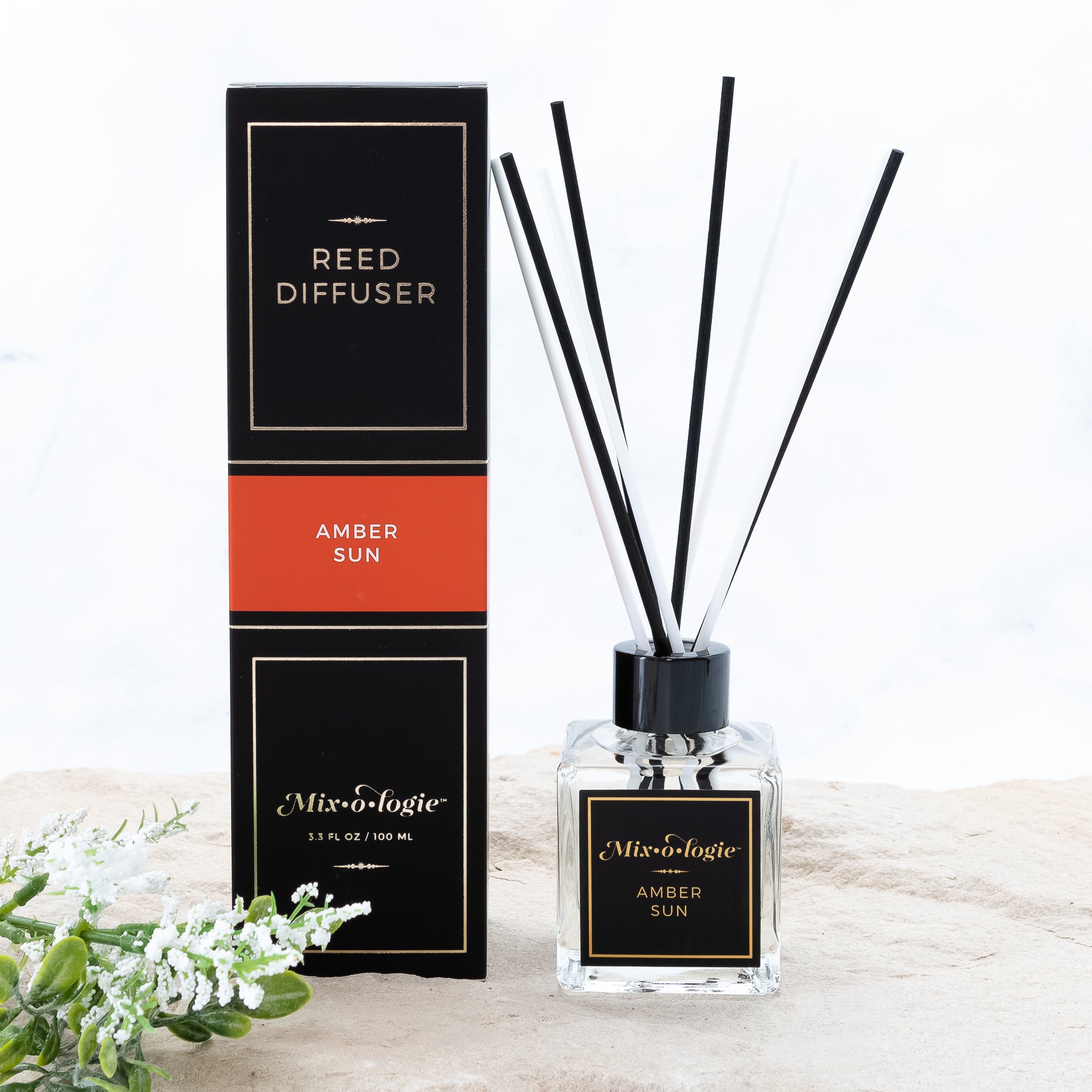 Amber Sun Reed Diffuser is a clear square glass container with black label that says Mixologie – Amber Sun. Has a black top and 4 white & 4 black reed sticks coming out of top, is 3.3 fl oz or 100 mL of clear scented liquid. Black rectangle package box with orange label. Box and diffuser are pictured with sand and greenery with white flowers.