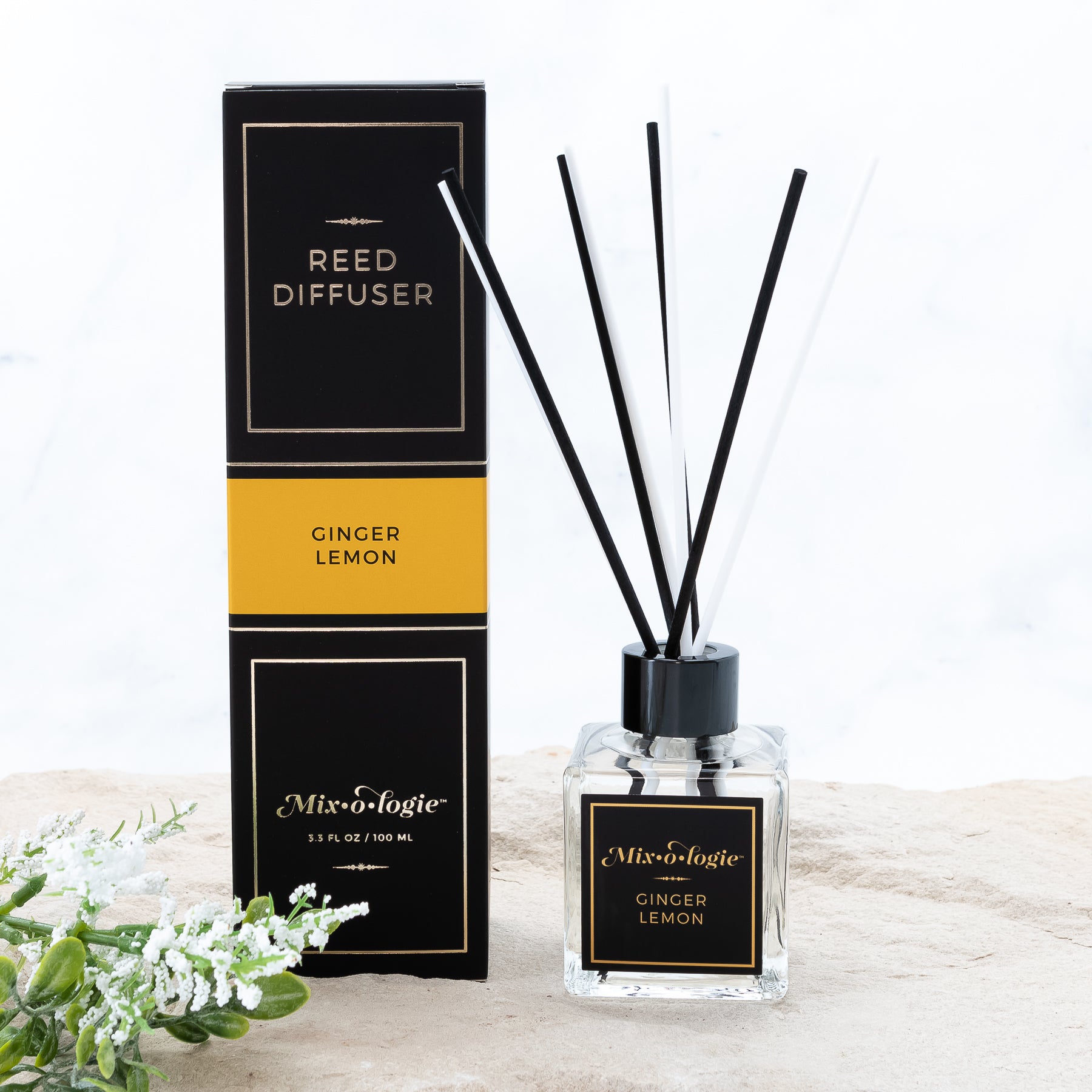 Ginger Lemon Reed Diffuser is a clear square glass container with black label that says Mixologie – Ginger Lemon. Has a black top and 4 white & 4 black reed sticks coming out of top, is 3.3 fl oz or 100 mL of clear scented liquid. Black rectangle package box with mustard label. Box and diffuser are pictured with sand and greenery with white flowers.
