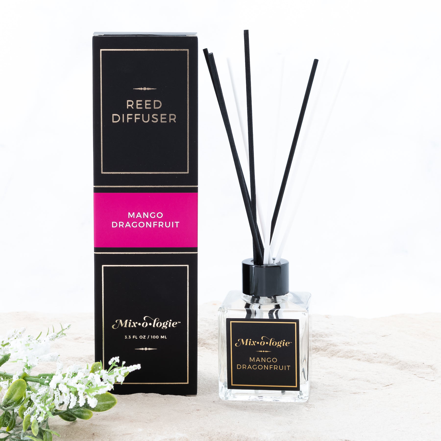 Mango Dragonfruit Reed Diffuser is a clear square glass container with black label that says Mixologie – Mango Dragonfruit. Has a black top and 4 white & 4 black reed sticks coming out of top, is 3.3 fl oz or 100 mL of clear scented liquid. Black rectangle package box with bright pink label. Box and diffuser are pictured in sand with greenery with white flowers.