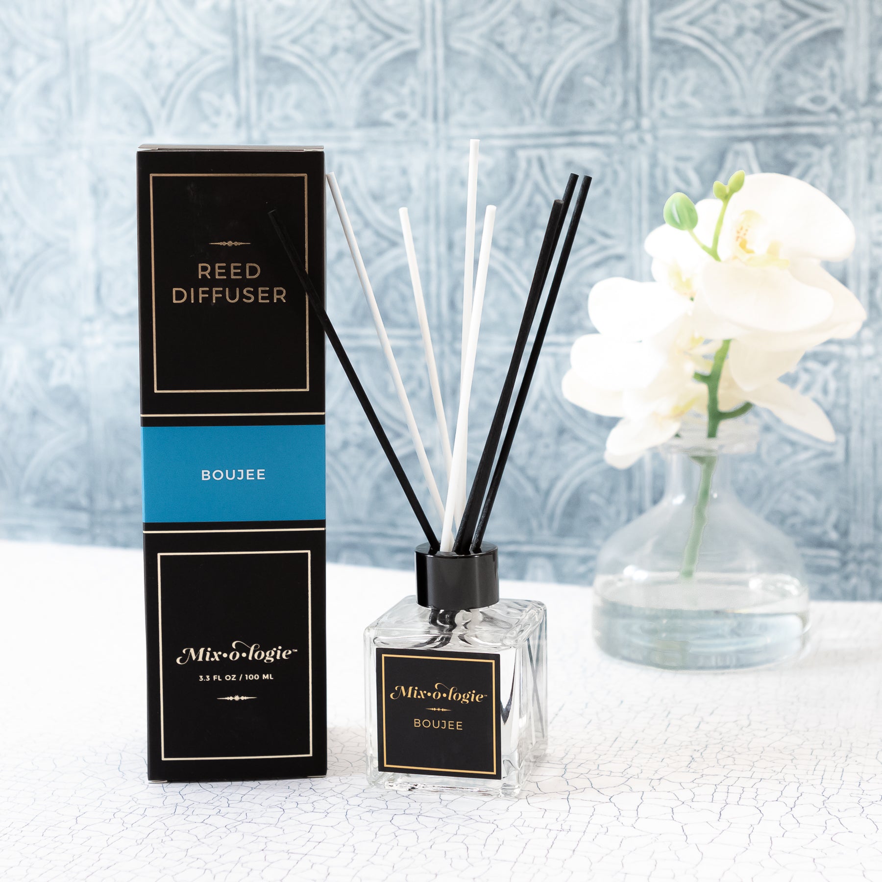 Boujee Reed Diffuser is a clear square glass container with black label that says Mixologie – Boujee. Has a black top and 4 white & 4 black reed sticks coming out of top, is 3.3 fl oz or 100 mL of clear scented liquid. Black rectangle package box with blue label. Box and diffuser are pictured on white countertop with clear vase and white flowers