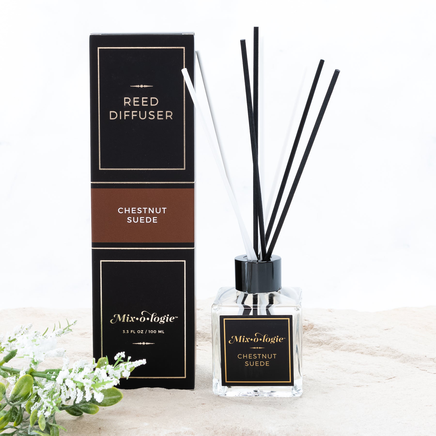 Chestnut Suede Reed Diffuser is a clear square glass container with black label that says Mixologie – Chestnut Suede. Has a black top and 4 white & 4 black reed sticks coming out of top, is 3.3 fl oz or 100 mL of clear scented liquid. Black rectangle package box with brown label. Box and diffuser are pictured with sand and greenery with white flowers.