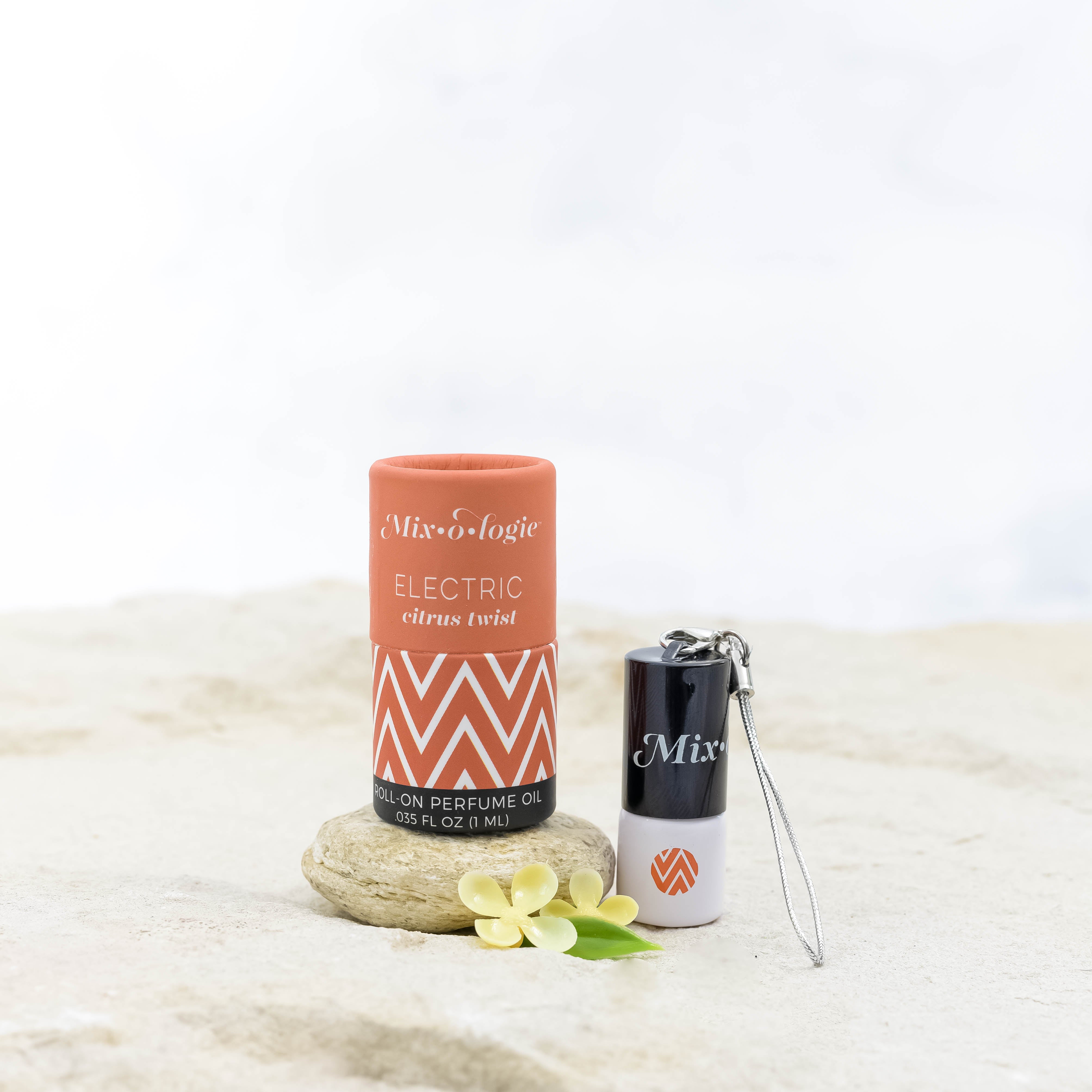 Electric (Citrus Twist) mini white cylinder rollerballs with black top and keychain attachment with orange chevron has .035 fl oz or 1 mL. Orange cylinder packing tube with orange chevron. Roll-on perfume oil. Mini rollerball and cylinder tube are in sand on a rock with yellow flowers.