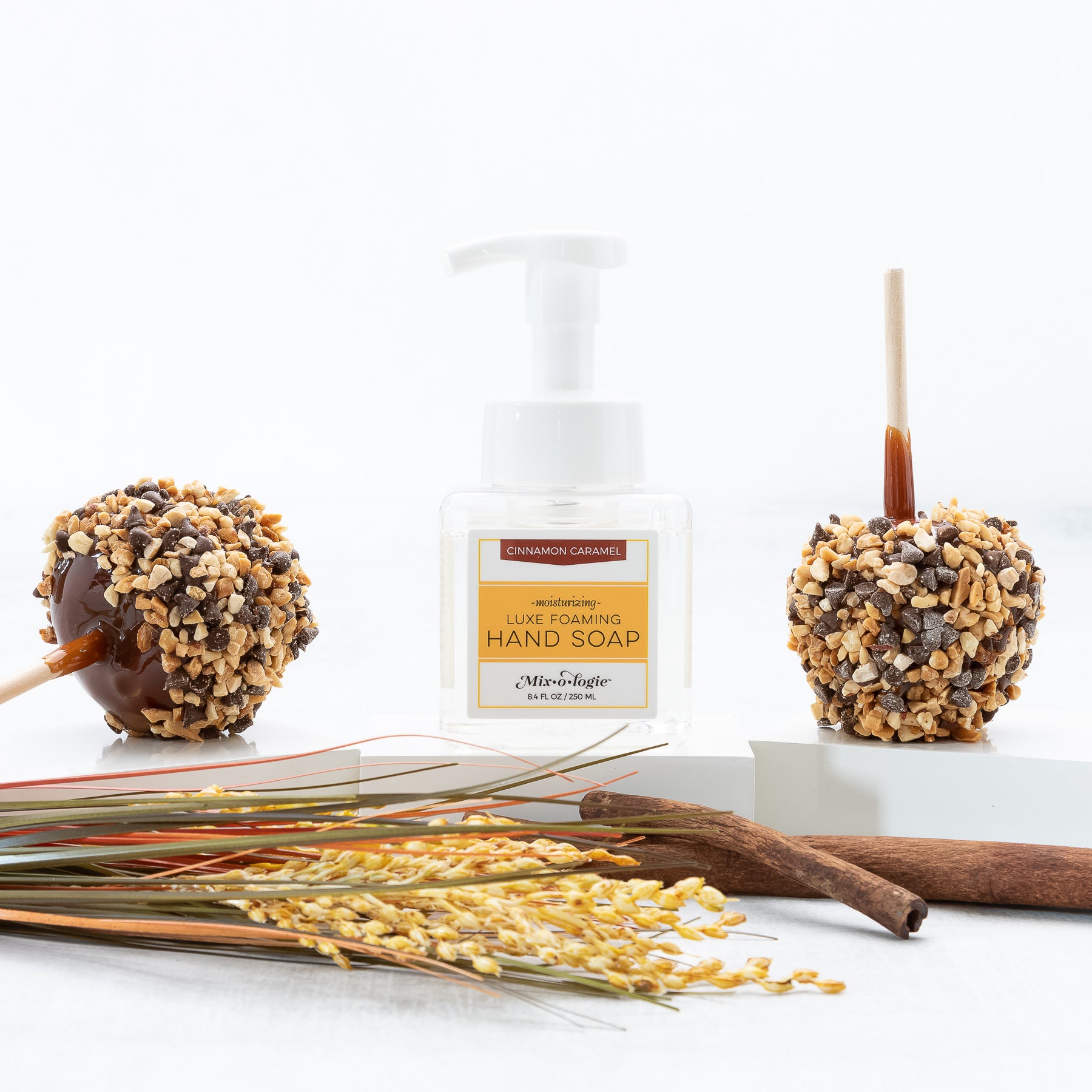 Moisturizing Luxe Foaming Hand Soap in Mixologie’s Cinnamon Carmel scent. Clear glass square shaped container with white pump that contains 8.4 fl oz or 250 ML of clear liquid. Pictured with Carmel apples, sticks, and greenery.