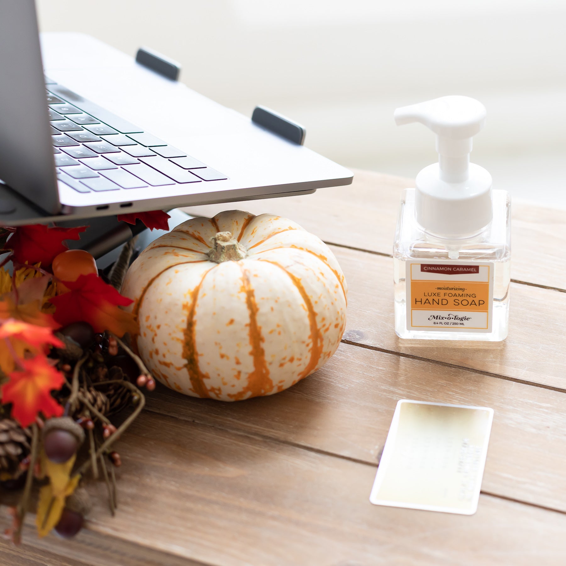 Moisturizing Luxe Foaming Hand Soap in Mixologie’s Cinnamon Carmel scent. Clear glass square shaped container with white pump that contains 8.4 fl oz or 250 ML of clear liquid. Pictured with fall leaves and pumpkin on table.