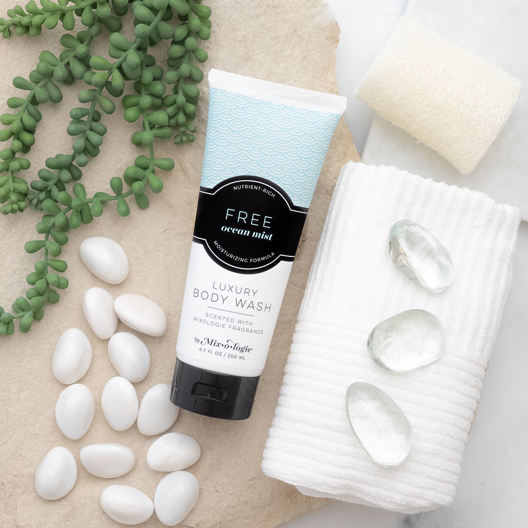 Luxury Body Wash in Mixologie’s Free (Ocean Mist) in light blue color sample package with black label. Contains 6.7 fl oz or 200 mL pictured on a large rock with greenery, white pebbles, a towel, and sponge.