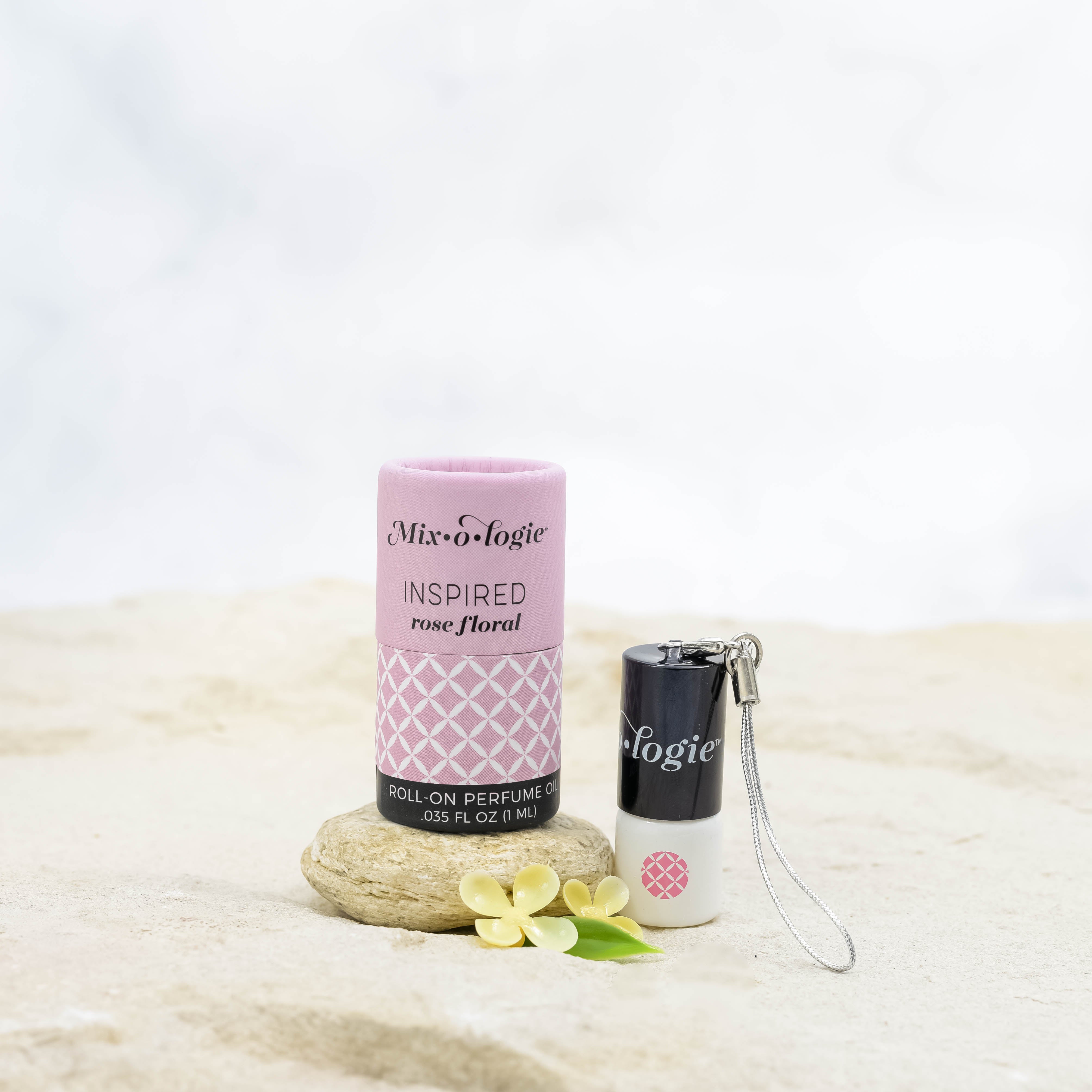 Inspired (Rose Floral) mini white cylinder rollerballs with black top and keychain attachment with pale pink diamond-like pattern and has .035 fl oz or 1 mL. Pale pink cylinder packing tube with pale pink wave-like pattern. Roll-on perfume oil. Mini rollerball and cylinder tube are in sand on a rock with yellow flowers.