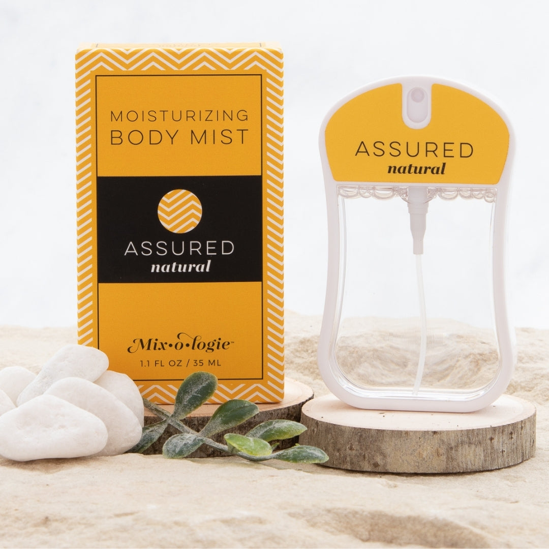  Assured (Natura) Moisturizing Body Mist in mustard color box and spray bottle with mustard color label and clear liquid. Spray bottle has 1.1 fl oz or 35 mL. Box and spray bottle are on wood in sand with greenery and pebbles.