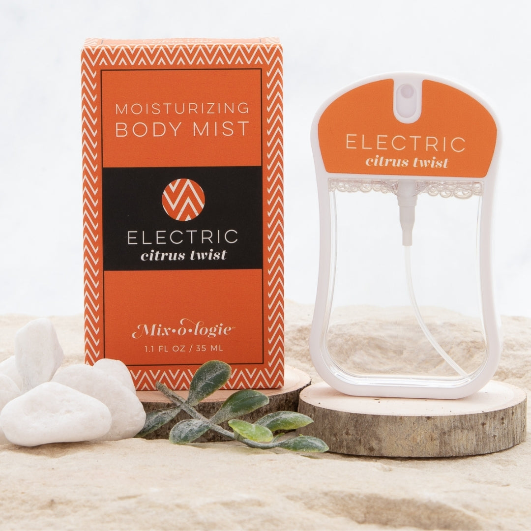 Electric (Citrus Twist) Moisturizing Body Mist in orange color box and rounded rectangle spray bottle with orange color label and clear liquid. Spray bottle has 1.1 fl oz or 35 ML. Spray bottle and box are pictured in the sand on wood with pebbles and greenery.