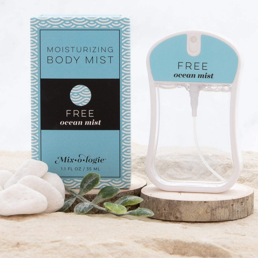 Free (Ocean Mist) Moisturizing Body Mist in pale blue color box and rounded rectangle spray bottle with pale blue color label and clear liquid. Spray bottle has 1.1 fl oz or 35 ML. Spray bottle and box are pictured in the sand on wood with pebbles and greenery.