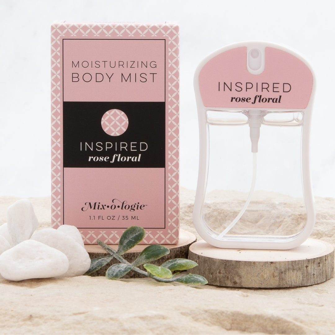 Inspired (Rose Floral) Moisturizing Body Mist in pale pink color box and rounded rectangle spray bottle with pale pink color label and clear liquid. Spray bottle has 1.1 fl oz or 35 ML. Spray bottle and box are pictured in the sand on wood with pebbles and greenery.