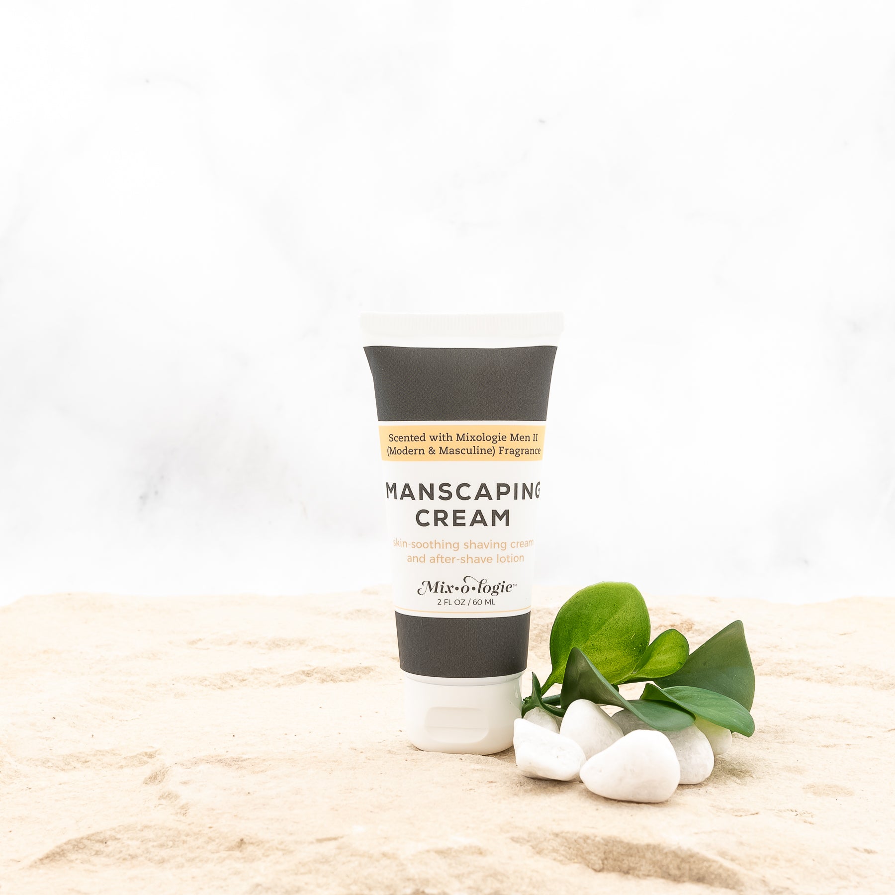 Men’s Manscaping Cream in Men’s II (Modern & Masculine) in a black and white tube with yellow accents. Skin-soothing shaving cream and after-shave lotion. 2 fl oz or 60 mL. Pictured in sand with rocks and greenery.