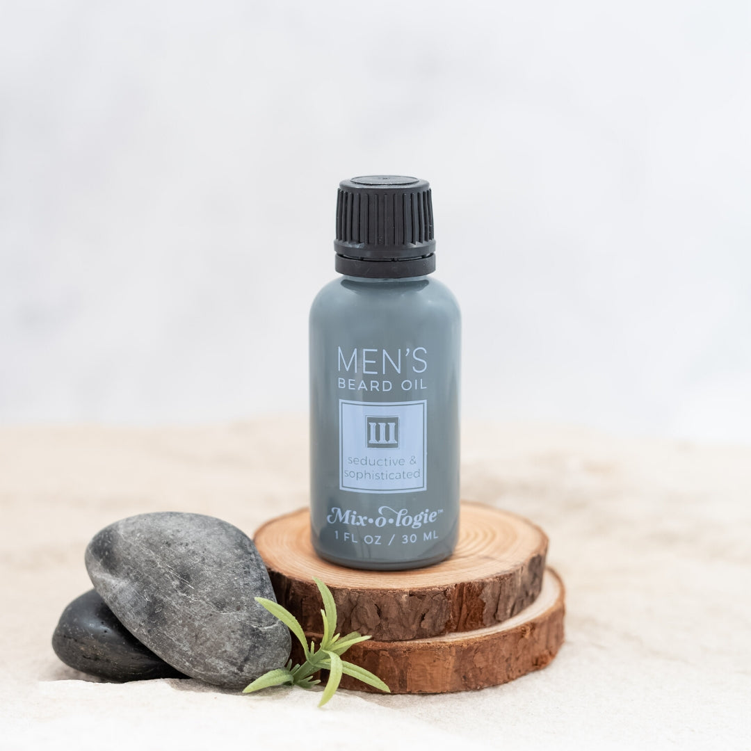 Men’s Beard Oil in Men’s III (Seductive & Sophisticated) in a black and grey bottle with blue colored accents. 1 fl oz or 30 mL. Pictured in sand on wood with rocks and greenery.