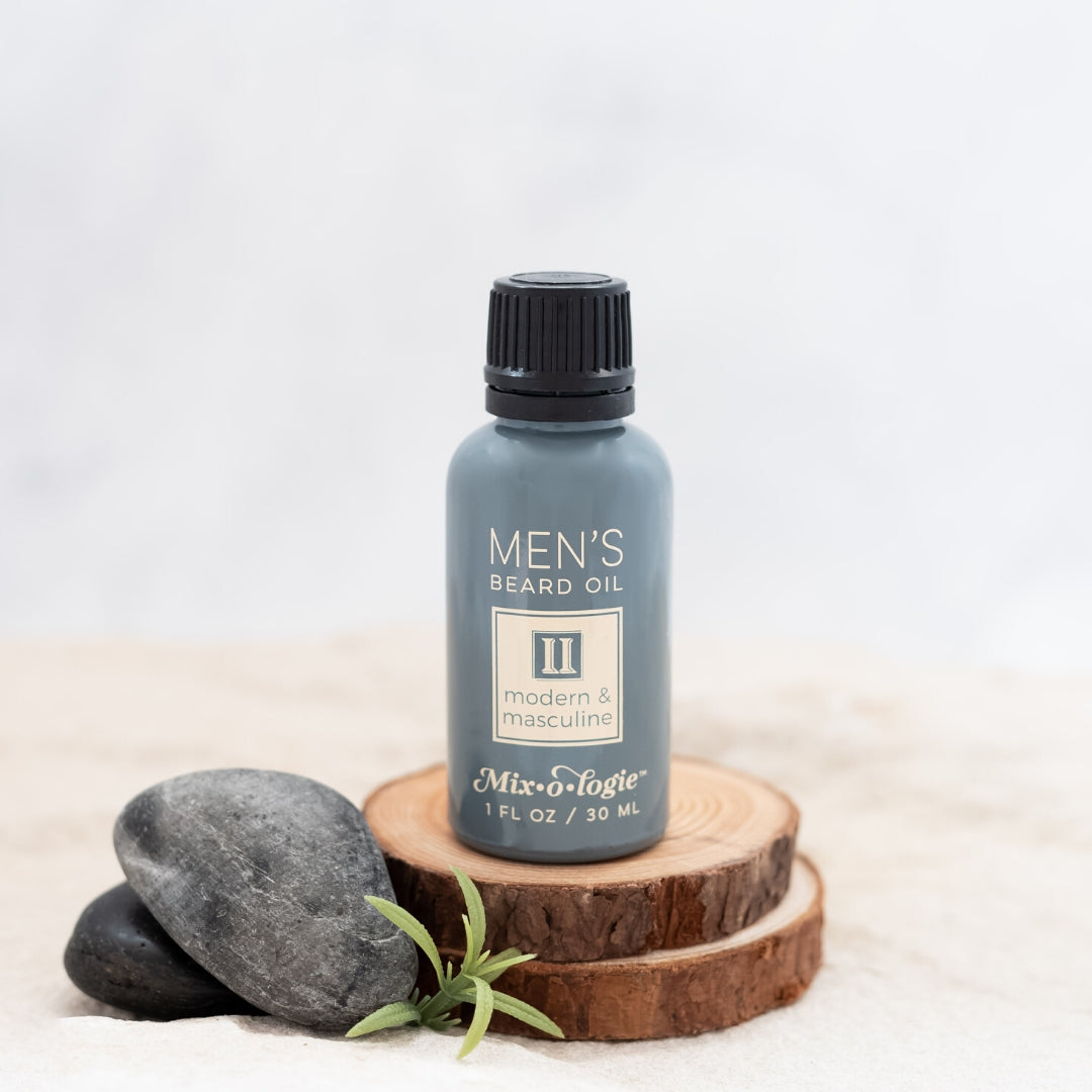 Men’s Beard Oil in Men’s II (Modern & Masculine) in a black and grey tube with yellow accents. 1 fl oz or 30 mL. Pictured in sand on wood with rocks and greenery.