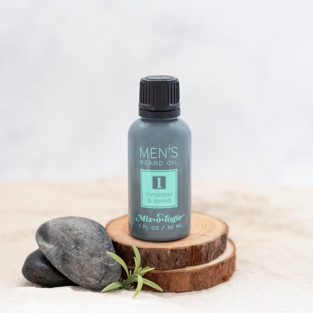Men’s Beard Oil in Men’s I (Timeless & Torrid) in a black and grey tube with green accents. 1 fl oz or 30 mL. Pictured in sand with rocks, wood, and greenery.