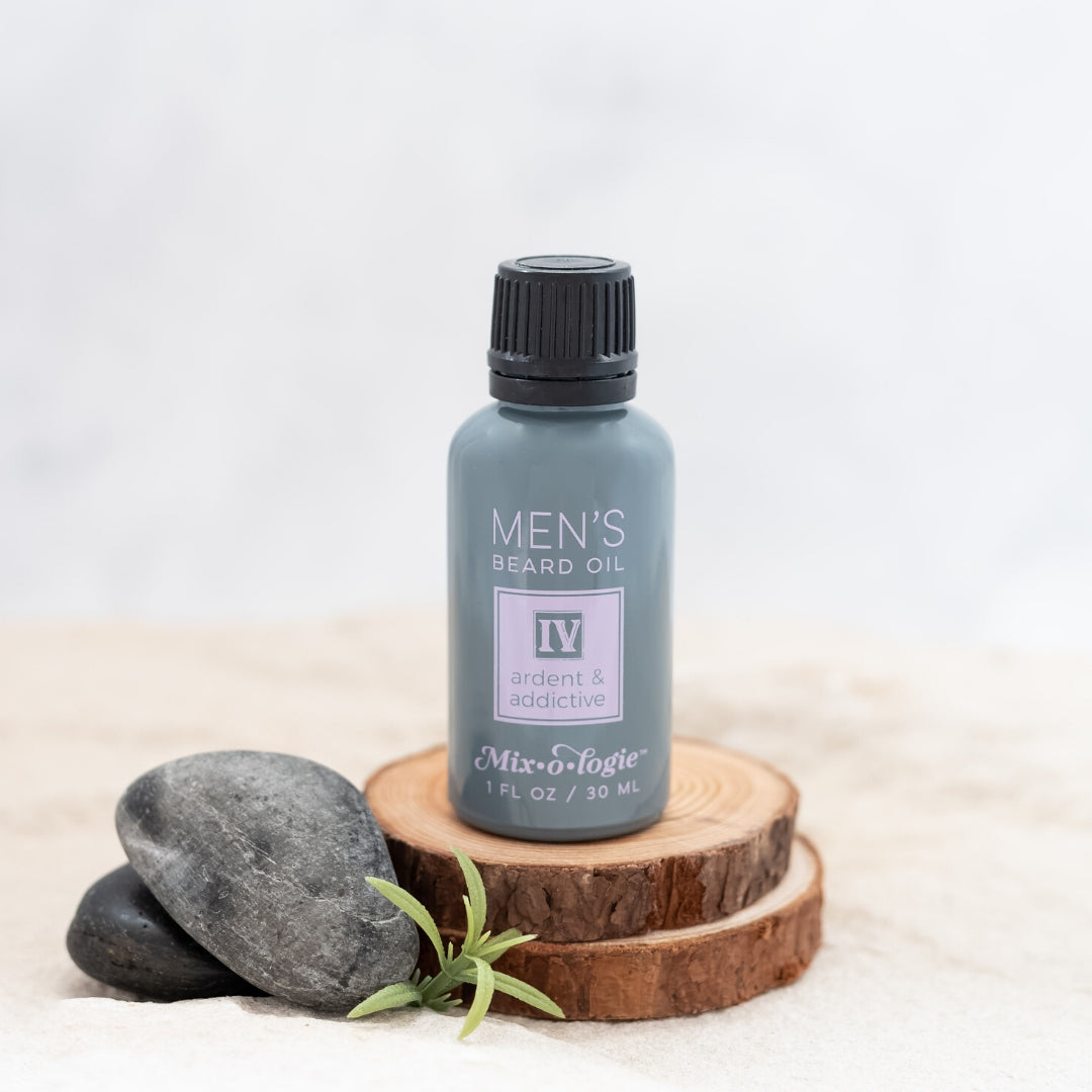 Men’s Beard Oil in Men’s IV (Ardent & Addictive) in a black and grey bottle with salmon-colored accents. 1 fl oz or 30 mL. Pictured on wood in sand with rocks and greenery.