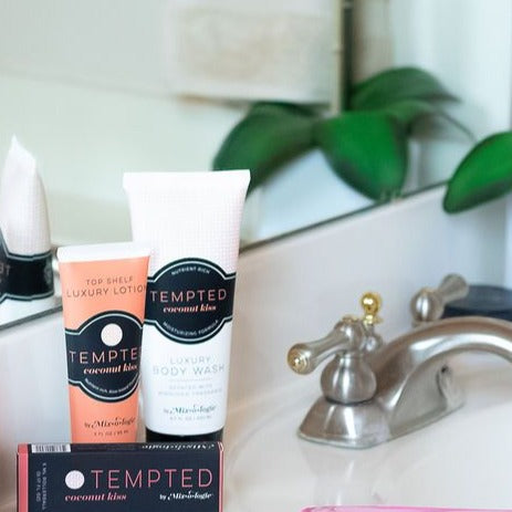 Luxury Body Wash in Mixologie’s Tempted (Coconut Kiss) in peach color sample package with black label. Contains 6.7 fl oz or 200 mL pictured on a bathroom counter with Tempted lotion and rollerball.