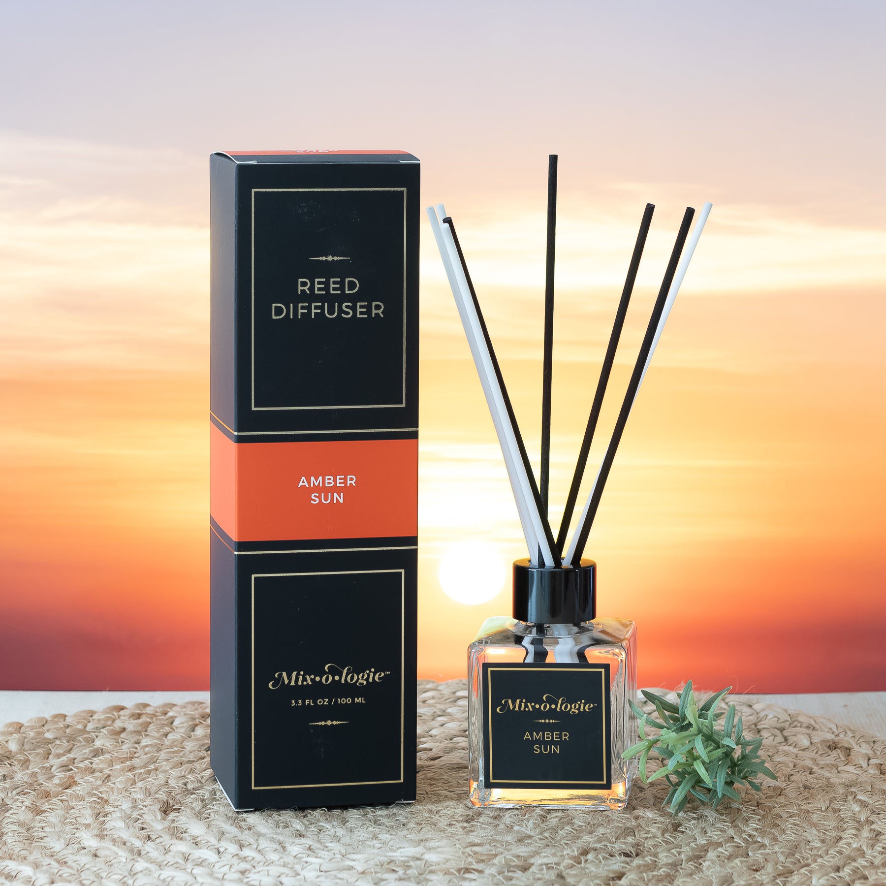 Amber Sun Reed Diffuser is a clear square glass container with black label that says Mixologie – Amber Sun. Has a black top and 4 white & 4 black reed sticks coming out of top, is 3.3 fl oz or 100 mL of clear scented liquid. Black rectangle package box with orange label. Box and diffuser are pictured with sunset background on woven place mat with greenery.