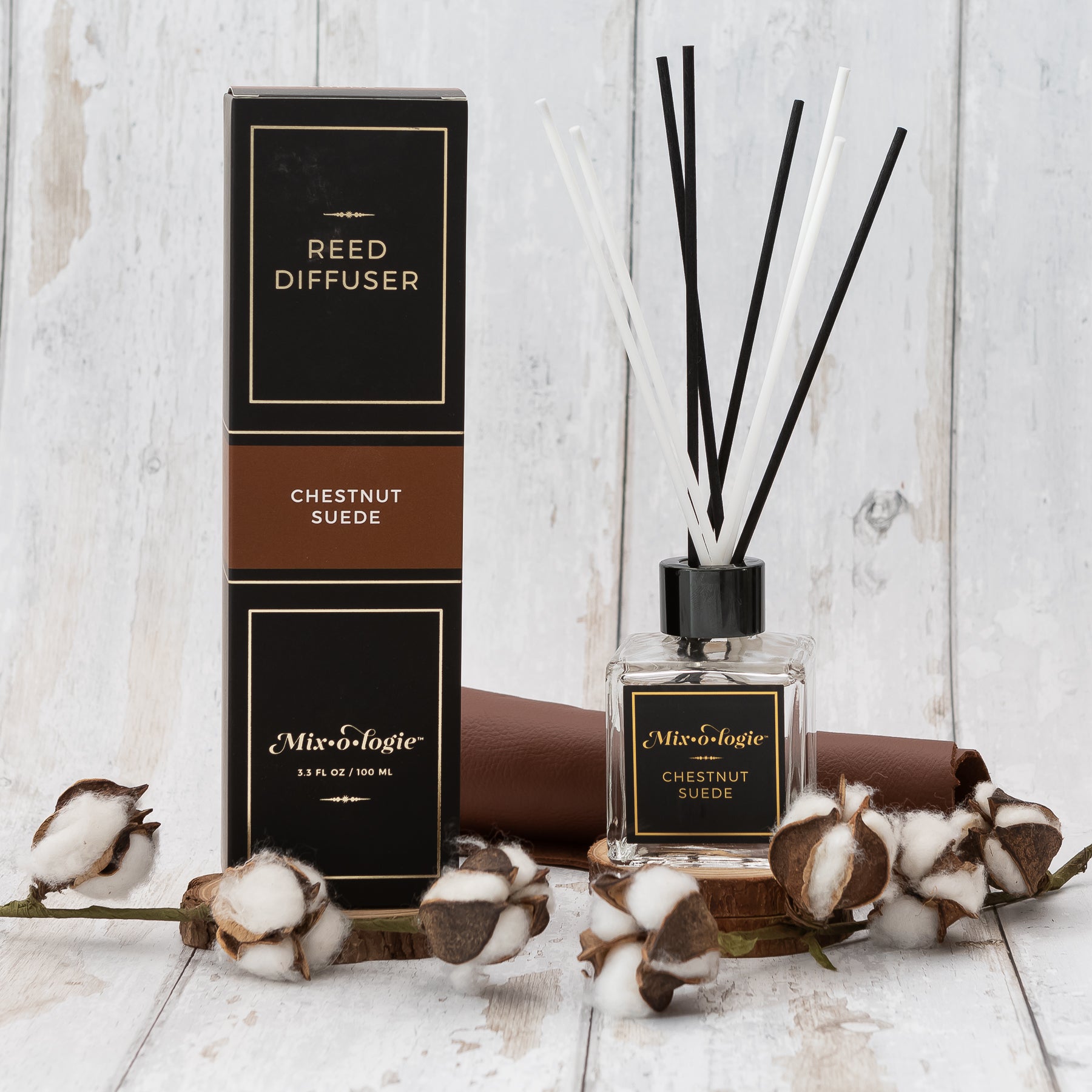 Chestnut Suede Reed Diffuser is a clear square glass container with black label that says Mixologie – Chestnut Suede. Has a black top and 4 white & 4 black reed sticks coming out of top, is 3.3 fl oz or 100 mL of clear scented liquid. Black rectangle package box with brown label. Box and diffuser are pictured with white wood and cotton. 