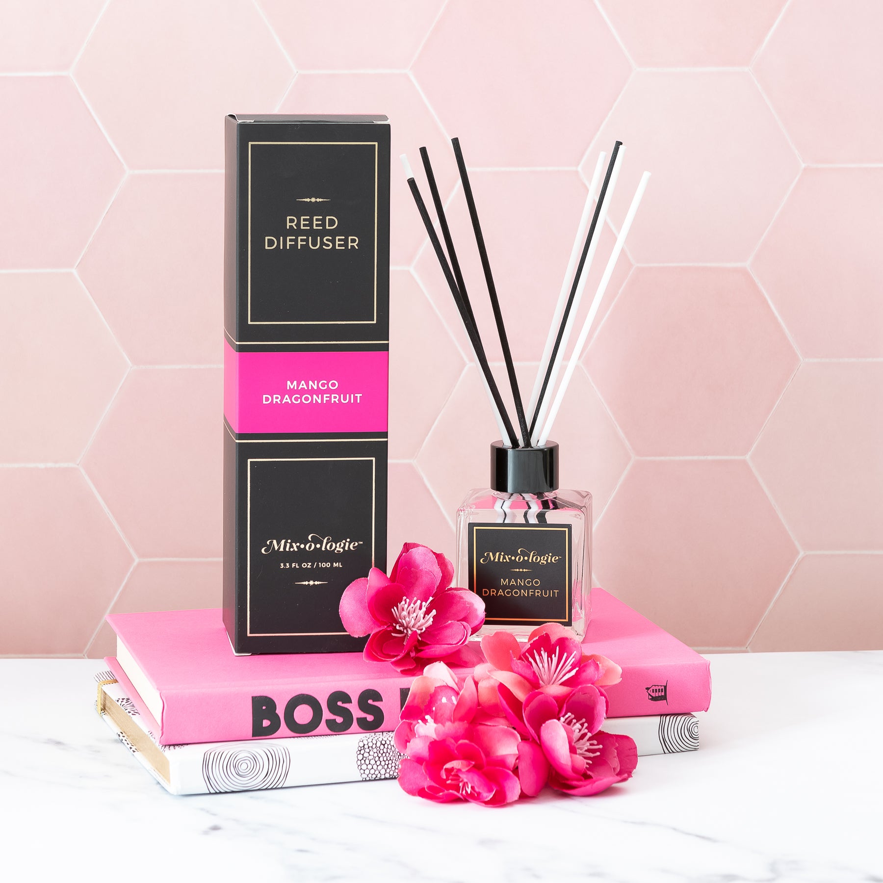 Mango Dragonfruit Reed Diffuser is a clear square glass container with black label that says Mixologie – Mango Dragonfruit. Has a black top and 4 white & 4 black reed sticks coming out of top, is 3.3 fl oz or 100 mL of clear scented liquid. Black rectangle package box with bright pink label. Box and diffuser are pictured pink tile background, books, and pink flowers.
