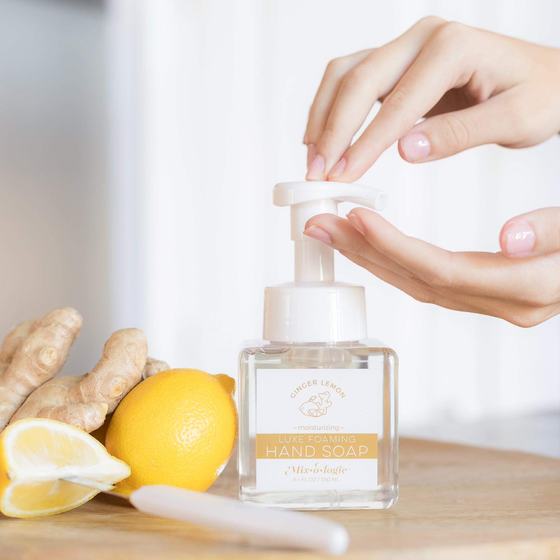 Moisturizing Luxe Foaming Hand Soap in Mixologie’s Ginger Lemon scent. Clear glass square shaped container with white pump that contains 8.4 fl oz or 250 ML of clear liquid. Pictured on a cutting board with lemons and ginger and a models hands.