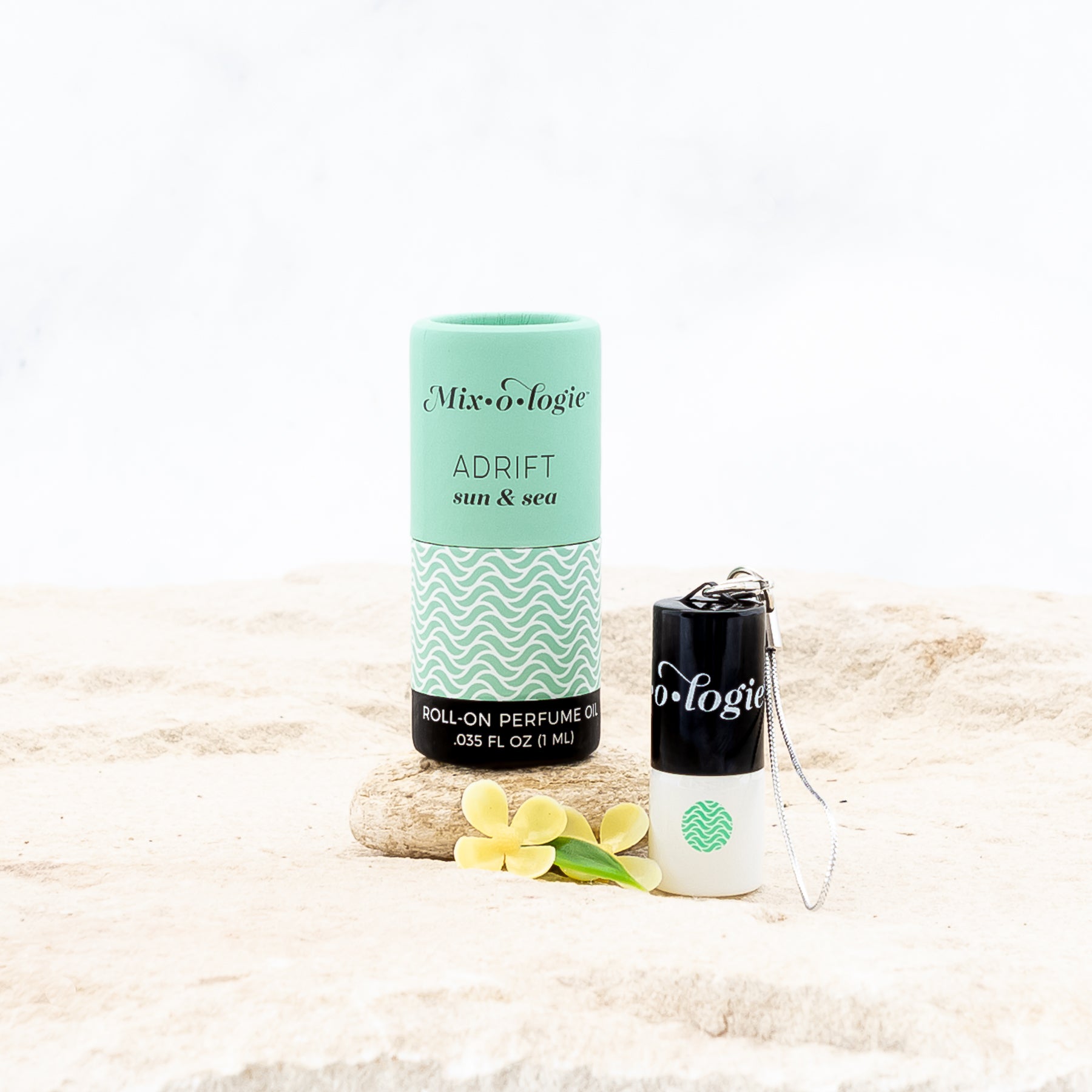 Adrift (Sun & Sea) mini white cylinder rollerballs with black top and keychain attachment with light green Adrift pattern has .035 fl oz or 1 mL. Light green cylinder packing tube. Mini rollerball and cylinder tube are in sand on a rock with yellow flowers.
