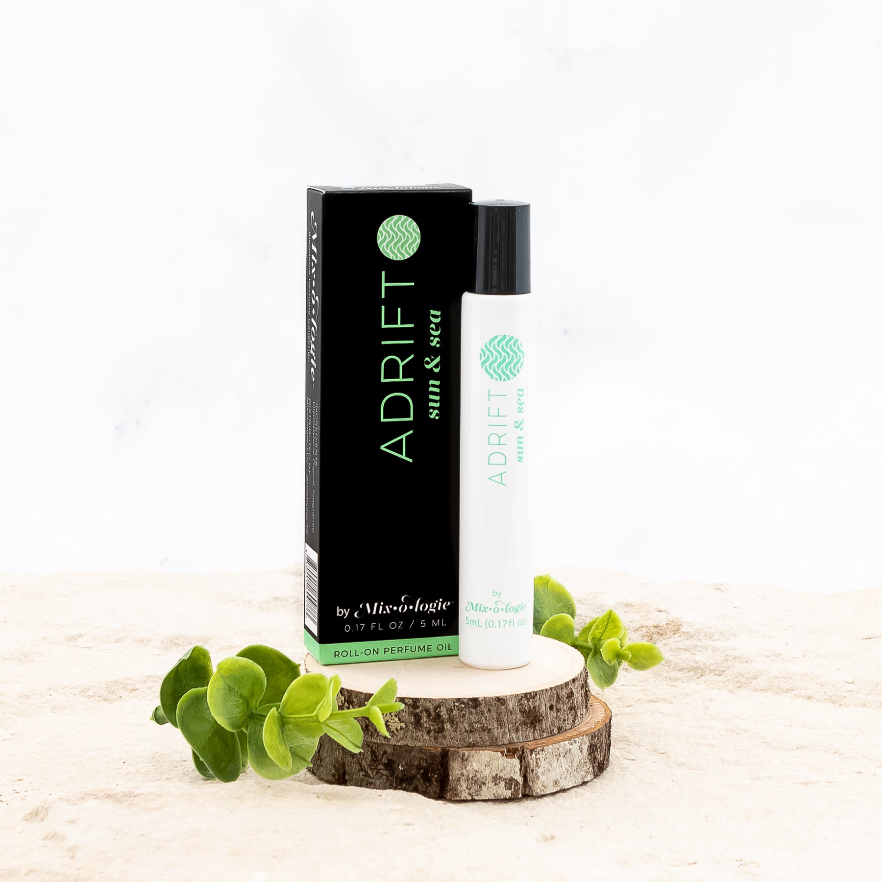 Adrift (Sun & Sea) White cylinder rollerball with light green color lettering with black box and light green color lettering. Rollerball has 0.17 fl oz or 5 mL. Rollerball and rollerball box are on wood in the sand with greenery.  