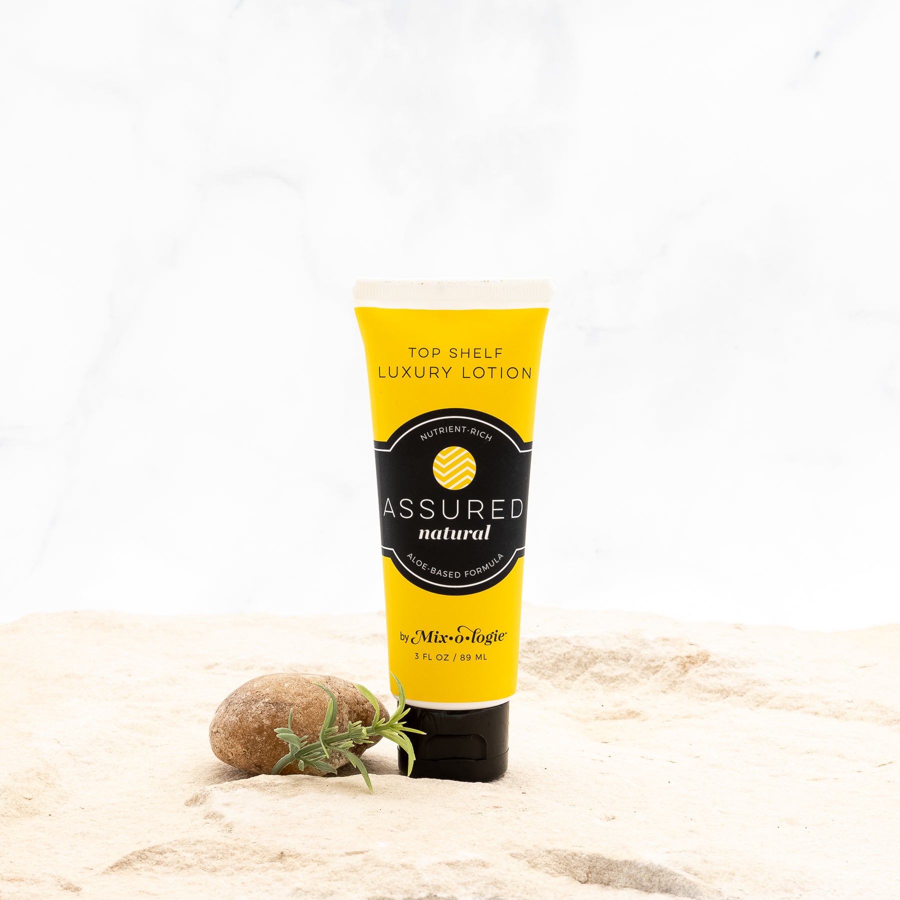 Assured (Natural) Top Shelf Lotion in mustard tube with black lid and label. Nutrient rich, aloe-based formula, tube has 5 fl oz or 89 mL. Pictured with white background, sand, rocks, and greenery. 