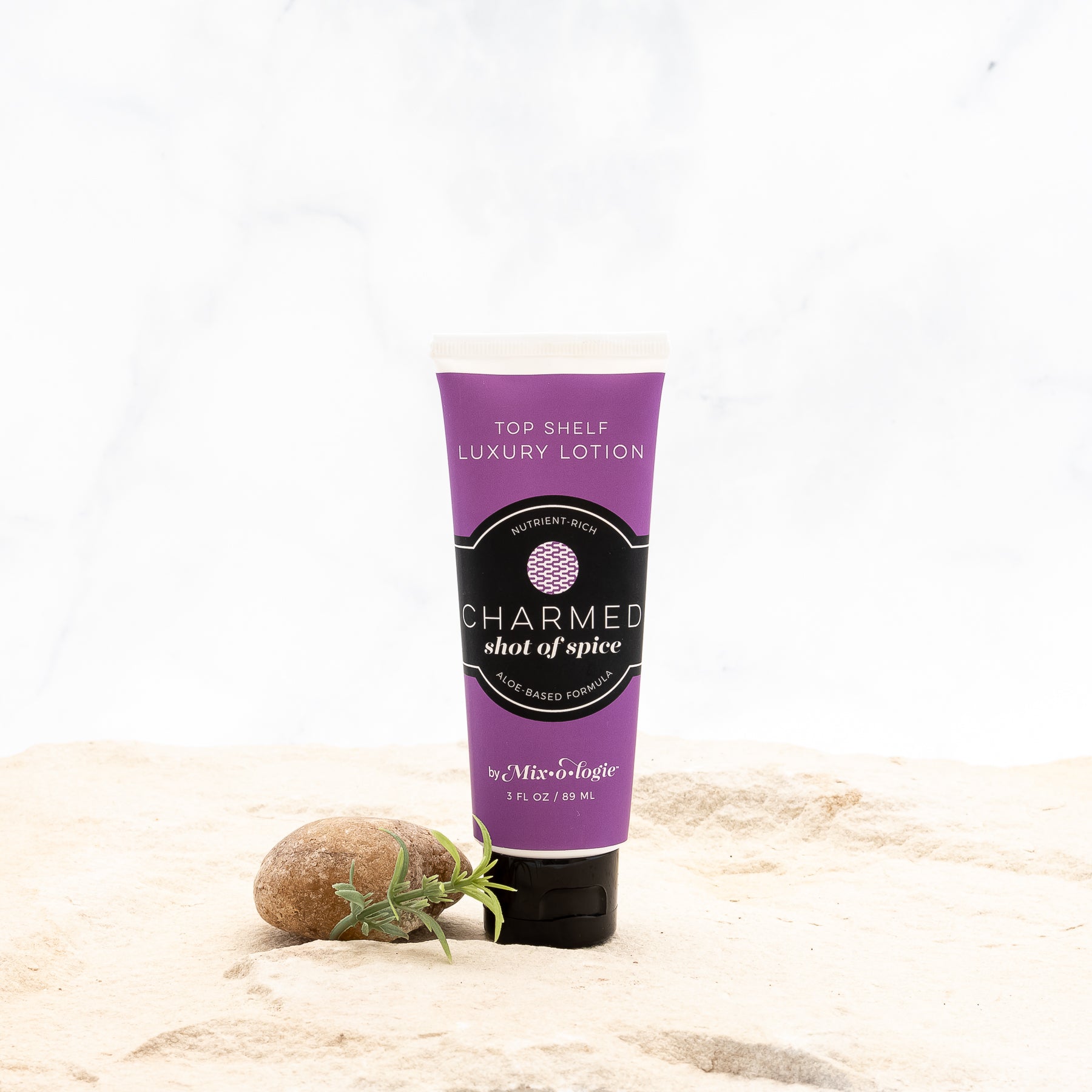 Charmed (Shot of Spice) Top Shelf Lotion in dark purple tube with black lid and label. Nutrient rich, aloe-based formula, tube has 5 fl oz or 89 mL. Pictured with white background, sand, rocks, and greenery. 