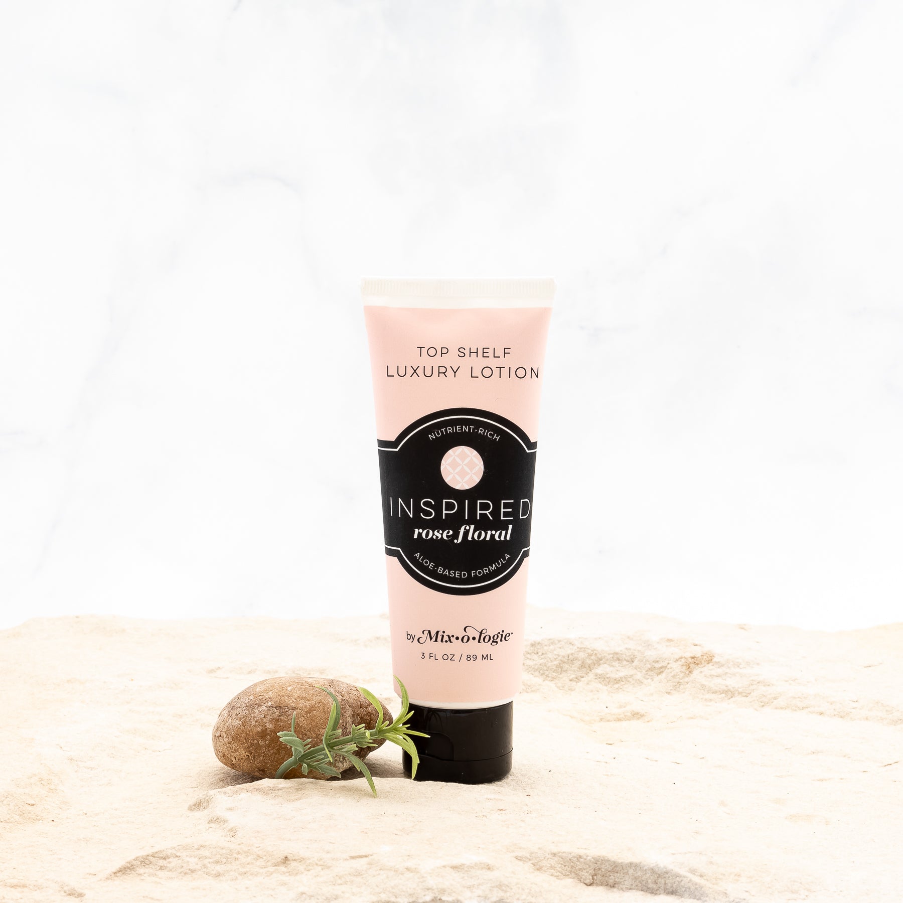 Inspired (Rose Floral) Top Shelf Lotion in pale pink tube with black lid and label. Nutrient rich, aloe-based formula, tube has 5 fl oz or 89 mL. Pictured with white background, sand, rocks, and greenery. 